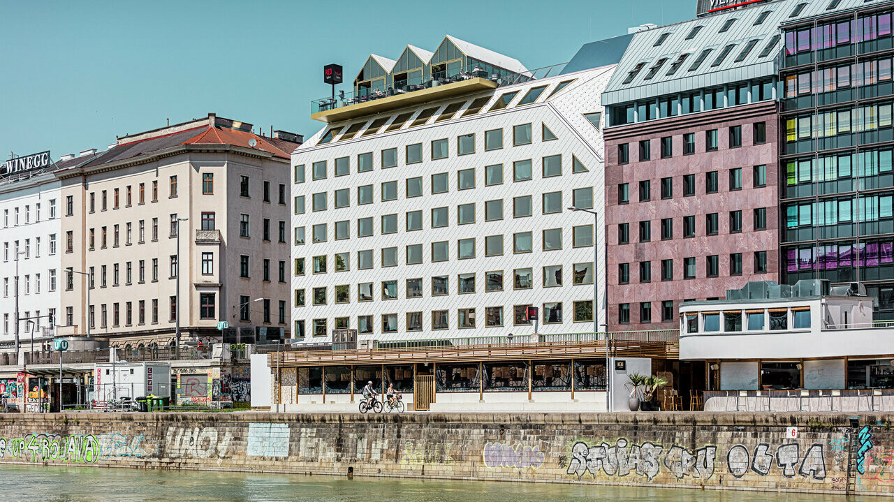 Vue sur les rives du canal du Danube à Vienne, avec une série de bâtiments d'architecture différente. Au premier plan, l'hôtel "THE ROCK Radisson RED Vienna", reconnaissable à sa façade blanche caractéristique en aluminium PREFA avec des losanges de toit et de façade 44 × 44 en P.10 blanc pur.
