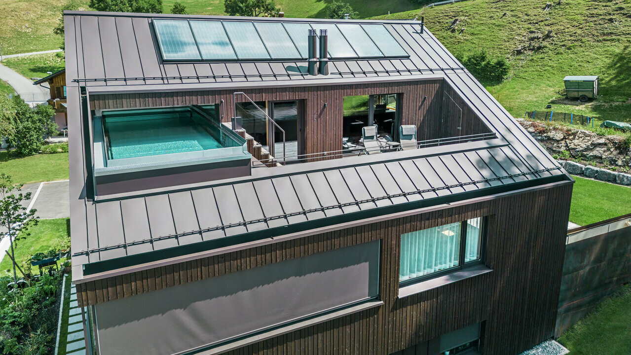Vue aérienne d'une maison moderne avec un toit en Prefalz PREFA brun noisette. Un toit-terrasse avec piscine offre un espace de détente exclusif avec vue sur le paysage vallonné et verdoyant.