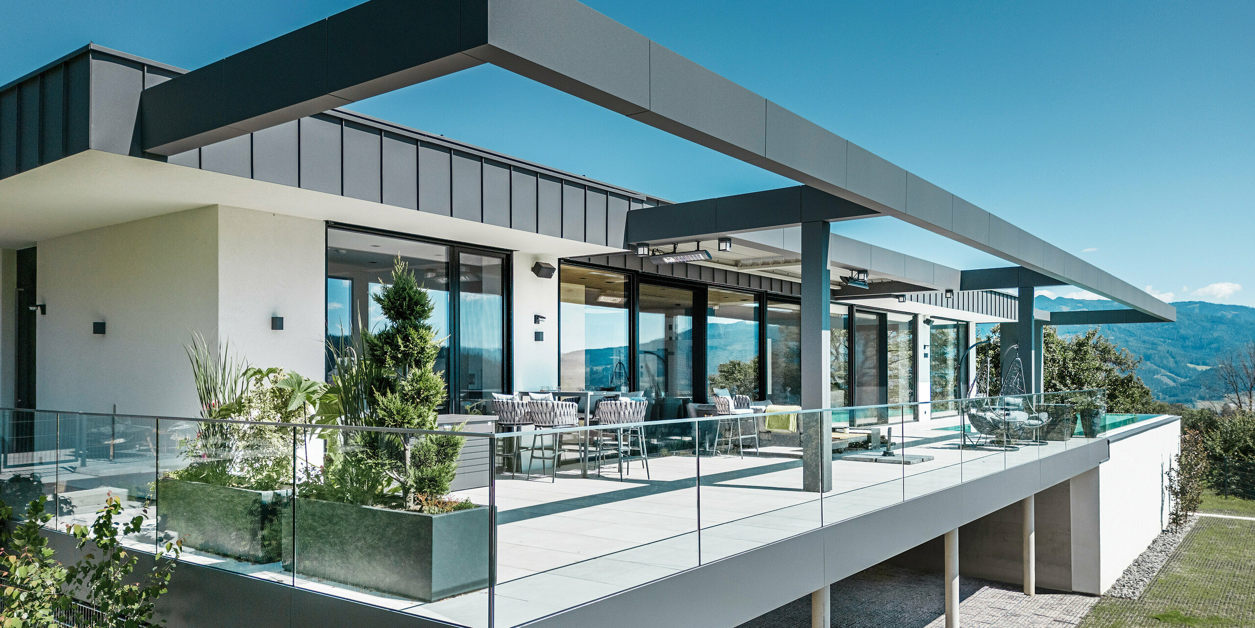 Luxuriöse Terrasse eines Einfamilienhauses in Pogier, Österreich mit eleganten Dach- und Fassadensystemen von PREFA. Die Kombination von PREFALZ und PREFABOND in dunkelgrau ergibt ein modernes und anspruchsvolles Design. Die großzügige Terrasse bietet einen atemberaubenden Ausblick und ist mit einem Glasgeländer ausgestattet, das eine offene und einladende Atmosphäre schafft. Der teilweise überdachte Außenbereich lässt die Schönheit der umliegenden Natur genießen und ist ideal zum Entspannen und geselligen Beisammensein.