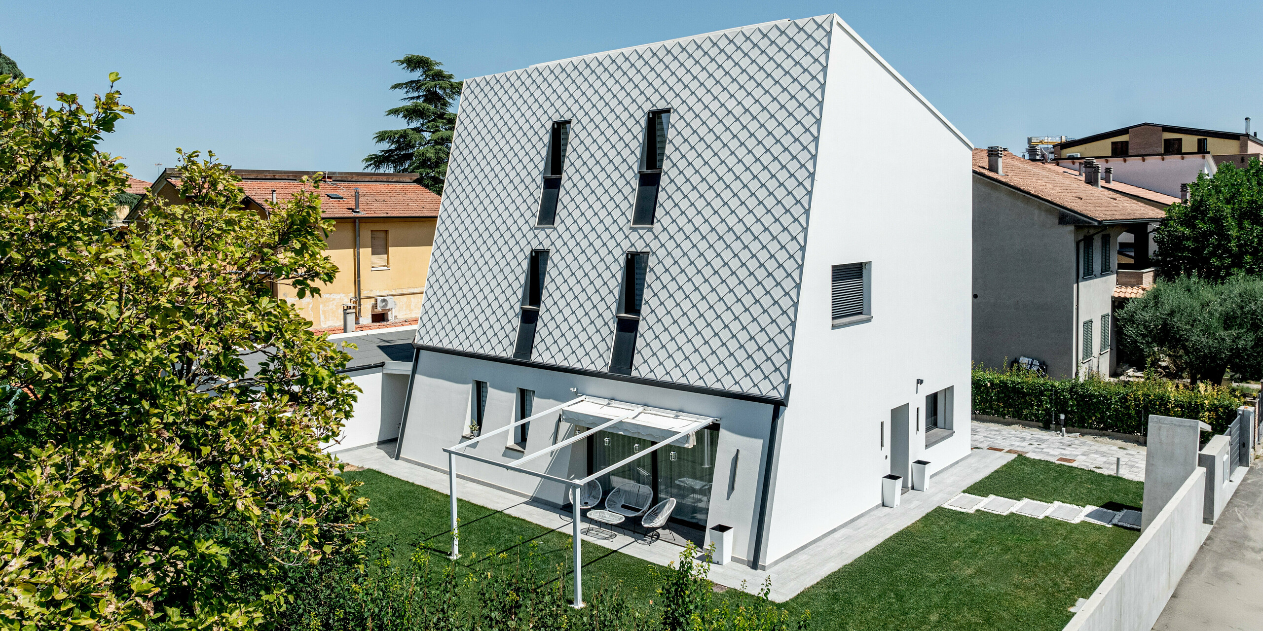 Schrägansicht eines modernen Einfamilienhauses in Forlì mit markanter, geneigter Hauptfassade aus PREFA Wandraute 29 × 29 in P.10 Reinweiß. Die diagonal angeordneten Aluminiumrauten bilden ein präzises, grafisches Muster, das im Sonnenlicht spannende Licht- und Schatteneffekte erzeugt. Schlanke, vertikale Fensteröffnungen durchbrechen die helle Oberfläche und betonen die Höhe des Baukörpers. Die langlebige PREFA Aluminiumfassade verbindet modernes Design mit hoher Witterungsbeständigkeit und minimalem Wartungsaufwand.