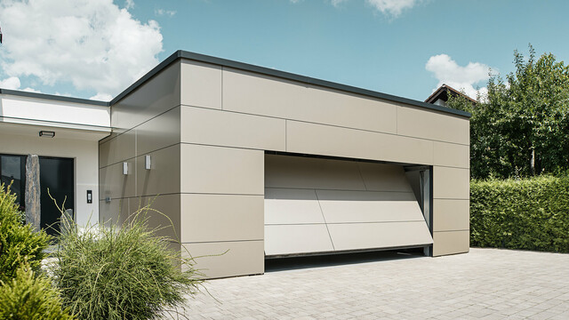 Gesamtansicht einer mit PREFABOND Aluminium Verbundplatten in Bronze verkleideten Garage. Das automatische Garagentor öffnet sich gerade und ist bereits ein Stück geöffnet. Direkt neben der Garage befindet sich der Hauseingang mit anthrazitfarbener Tür und weißer Putzfassade.