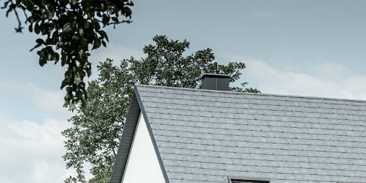 Il tetto a due falde della classica casa unifamiliare è stato rivestito con la nuova tegola R.16 PREFA in grigio pietra. Sulla superficie del tetto sono stati inseriti tre lucernari e sono stati montati i nasi paraneve. La facciata è di colore bianco semplice.