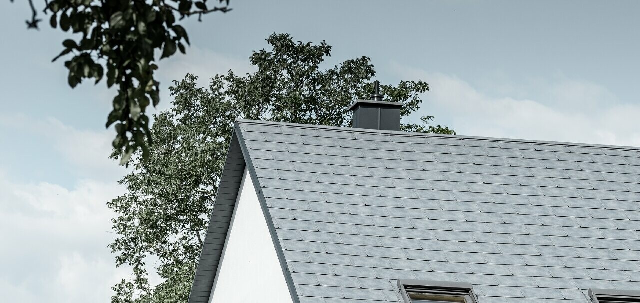 Le toit à deux pans de la maison individuelle classique a été recouvert avec la nouvelle tuile R.16 PREFA couleur gris pierre. Trois fenêtres de toit ont été intégrées dans la surface de toit et des arrêts neige ont été montés. La façade a été simplement laissée blanche.