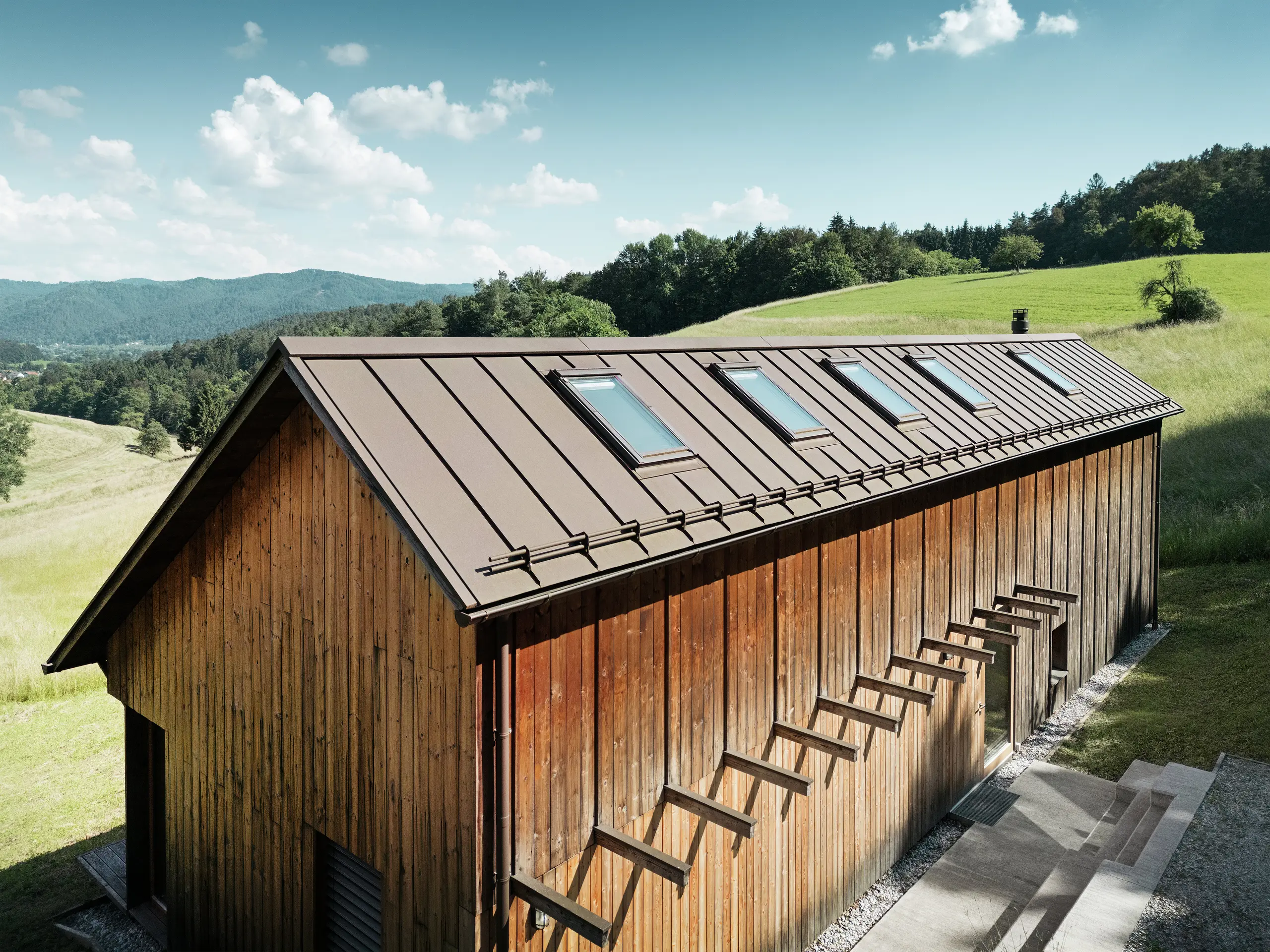 Casa in legno dalla struttura architettonica chiara con tetto in alluminio PREFALZ di alta qualità in P.10 marrone. Il tetto PREFA, che richiede poca manutenzione, convince per le sue linee precise, l'elevata resistenza ai raggi UV e alla corrosione e la perfetta integrazione delle finestre per tetti. La combinazione di una facciata in legno sostenibile e di un tetto doppia aggraffatura durevole si inserisce armoniosamente nell'idilliaco paesaggio collinare. Il tetto a falde asimmetriche è dotato di cinque lucernari posizionati con precisione che forniscono luce all'interno.