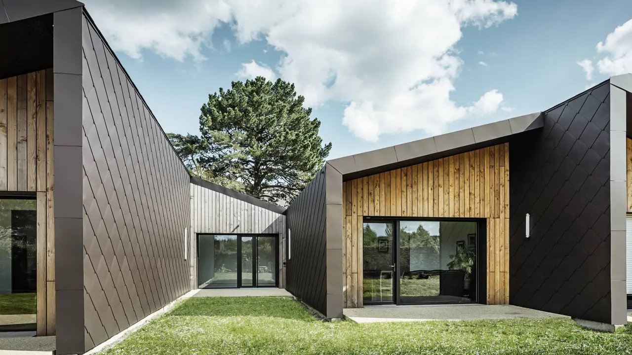 L'immagine mostra un'architettura moderna con una facciata composta da scaglie  44 × 44 PREFA per tetto e facciate nel colore marrone testa di moro. La struttura a diamante del rivestimento in alluminio conferisce all'edificio un aspetto elegante e geometrico. La facciata combina armoniosamente elementi in legno naturale e ampie superfici vetrate, creando un'atmosfera calda e invitante. Il prato verde e il cielo limpido rafforzano l'impressione di modernità e naturalezza dell'architettura.