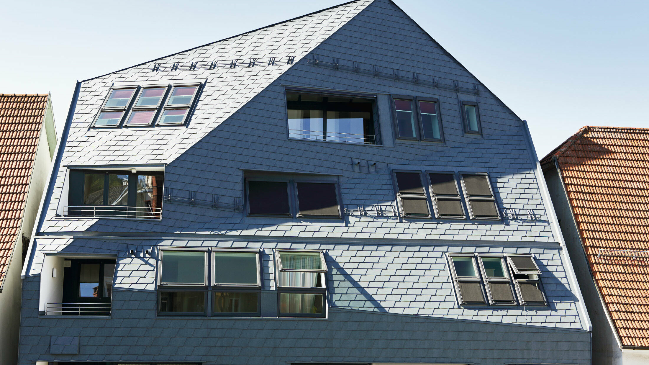 Maison galerie avec façade plissée, revêtue du bardeau de toiture et de façade PREFA en P.10 gris pierre avec de nombreuses fenêtres