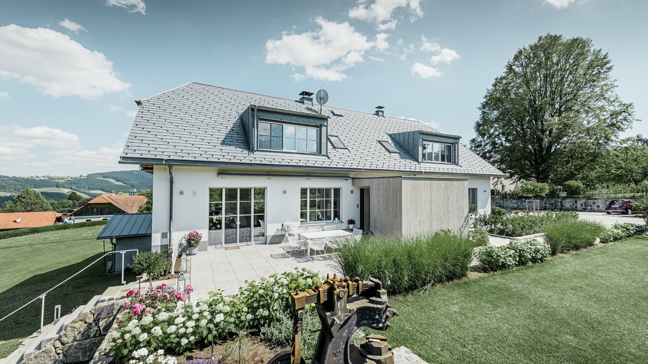 Maison individuelle classique avec toit en demi-croupe ; maison avec toit rénové à l'aide de bardeaux PREFA couleur gris pierre, avec beau jardin et grande terrasse.