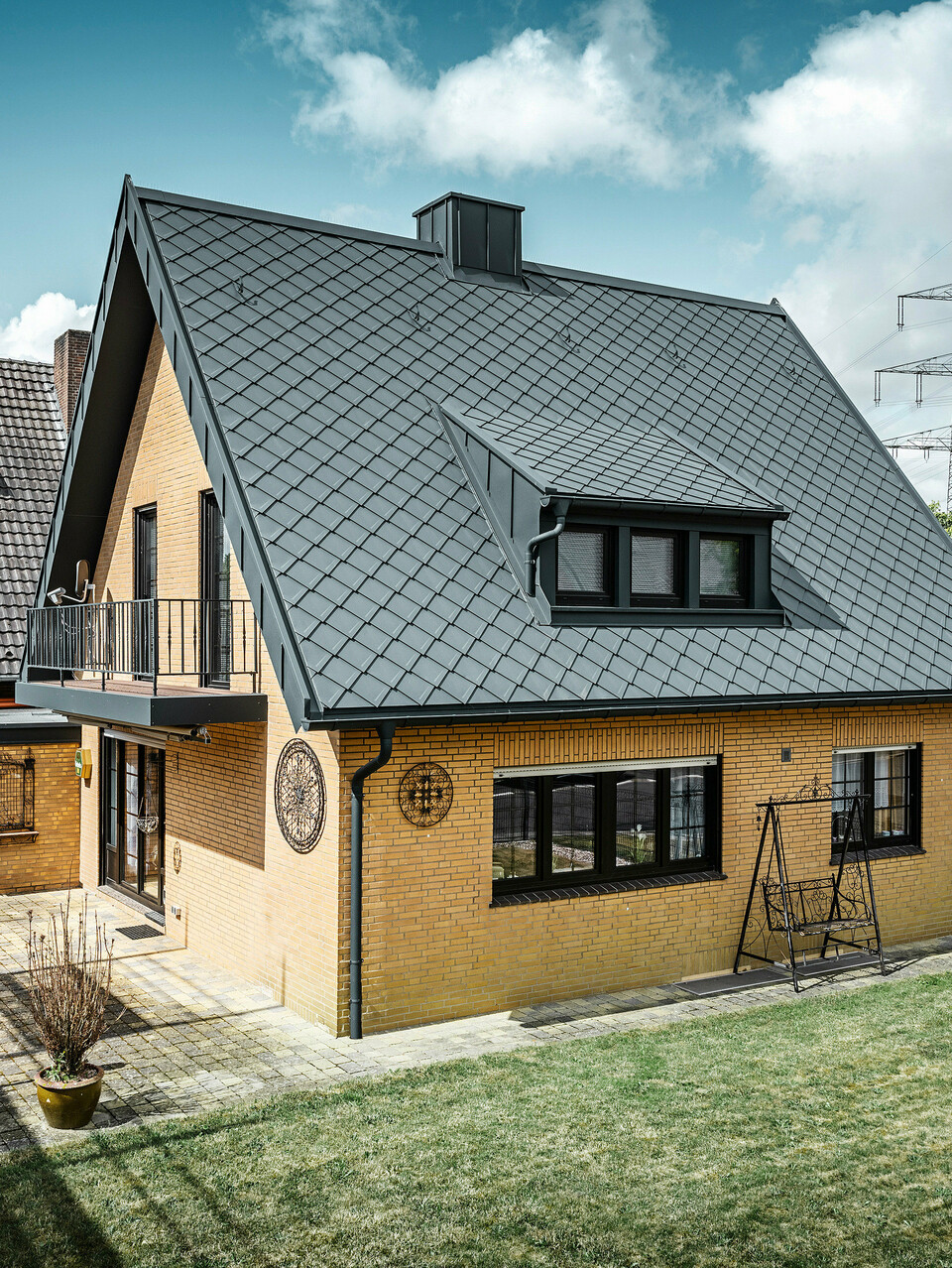 Ein Einfamilienhaus in Tönisvorst mit einem PREFA Dach aus Dachrauten 29×29 in P.10 Anthrazit, das ein herausstechendes Rautenmuster bietet. Schleppgauben, Kamin und der Eingangsbereich sind mit PREFALZ verkleidet, was zusammen mit Ablaufrohr und Hängerinne in P.10 Anthrazit dem Haus einen zeitlosen und eleganten Look verleiht. Das Backsteingebäude mit dem Aluminiumdach harmoniert mit der ruhigen, grünen Umgebung der Siedlung.