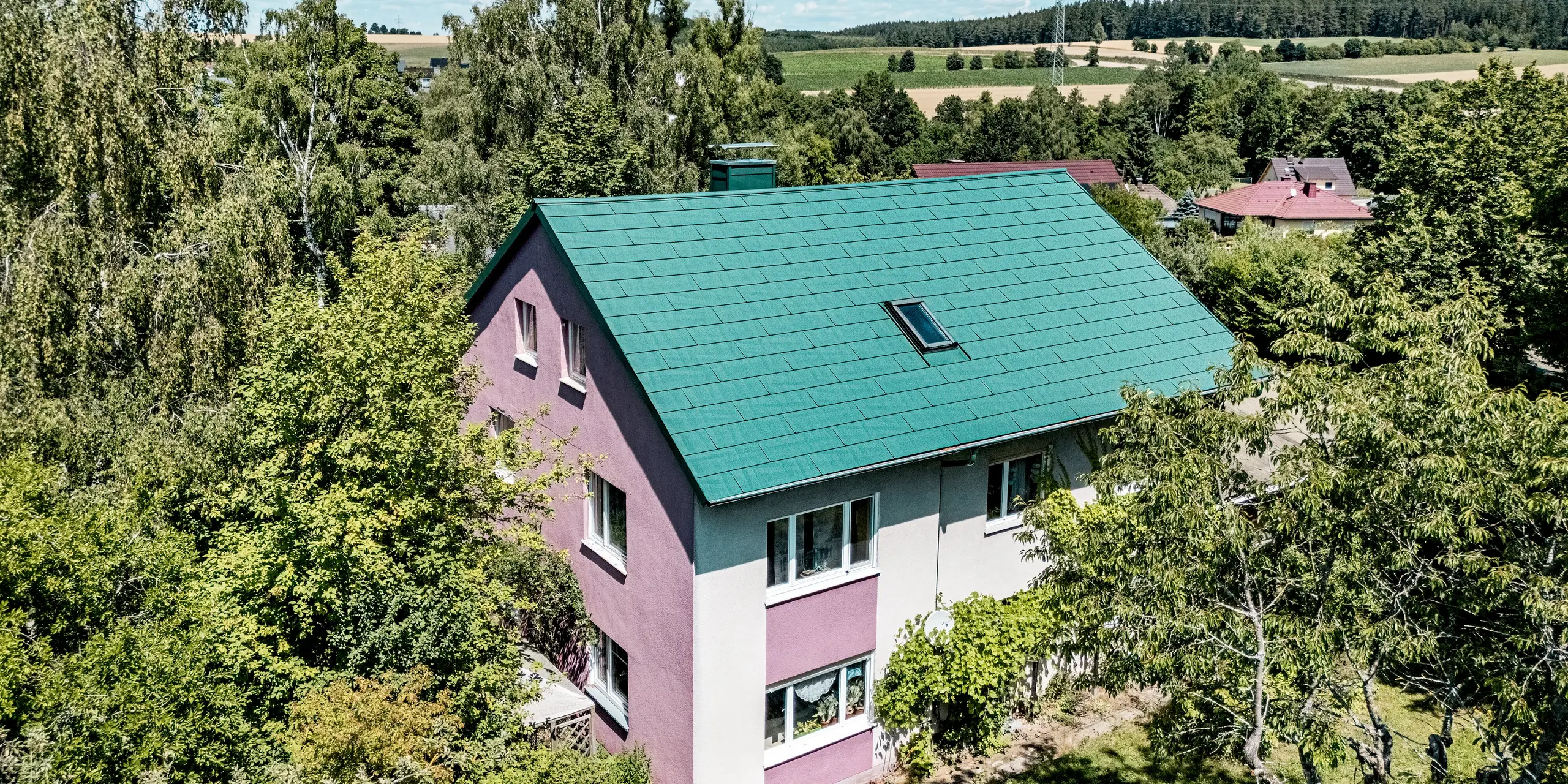 Sanierung eines Einfamilienhauses in Schwarzenbach-Martinlamitz mit PREFA Dachpaneel FX.12 in P.10 Moosgrün. Das Bild zeigt das modernisierte Aluminiumdach in kräftigem Moosgrün, das durch seine klare Linienführung und strukturierte Oberfläche auffällt. Der Kontrast zur violett-weißen Fassade verleiht dem Gebäude einen einzigartigen Charakter. Eingebettet in die grüne Landschaft wirkt das Dach modern und dennoch naturnah. Langlebig, sturmsicher und wartungsfrei – das PREFA Dach vereint Funktionalität mit stilvollem Design.
