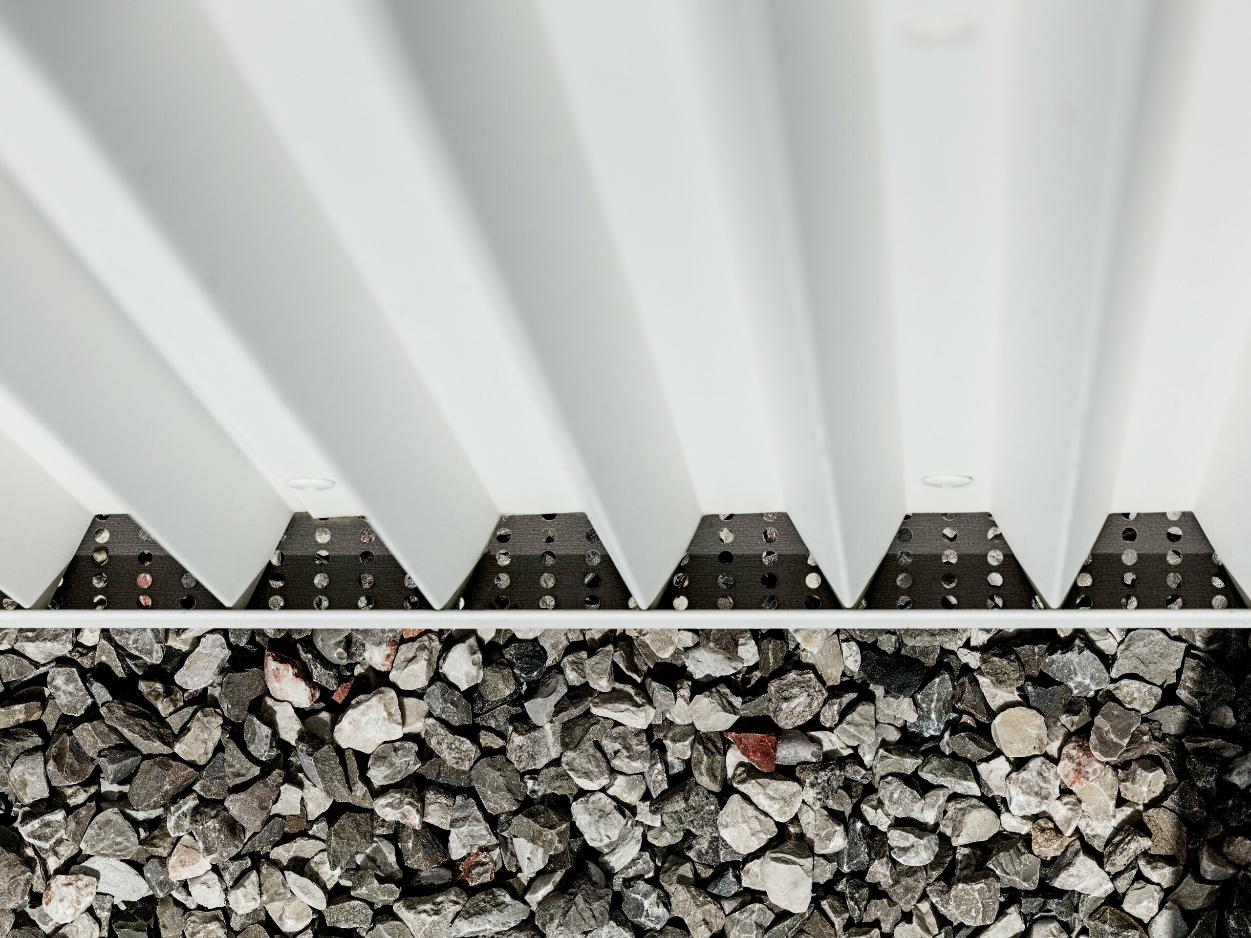 Das Bild zeigt einen Detailausschnitt der hinterlüfteten PREFALZ-Fassade aus reinweißem Aluminium, die mit präzise gefalteten Zickzack-Formen versehen ist. Diese innovative Falztechnik gewährleistet nicht nur eine optisch ansprechende Struktur, sondern auch eine hervorragende Belüftung und Witterungsbeständigkeit der Fassade. Die Verbindung der Fassadenelemente ist durch spezialisierte, unsichtbare Befestigungen realisiert, die eine besonders saubere und langlebige Oberfläche ermöglichen. Der untere Bereich des Bildes zeigt den Kiesbelag, der in der Umgebung des Gebäudes zur Drainage und Stabilisierung dient, was die Funktionalität der Fassadengestaltung weiter unterstreicht. Durch die Verwendung von PREFALZ werden ästhetische Ansprüche und technische Anforderungen gleichermaßen erfüllt, was das Material zu einer idealen Wahl für moderne und nachhaltige Architektur macht.