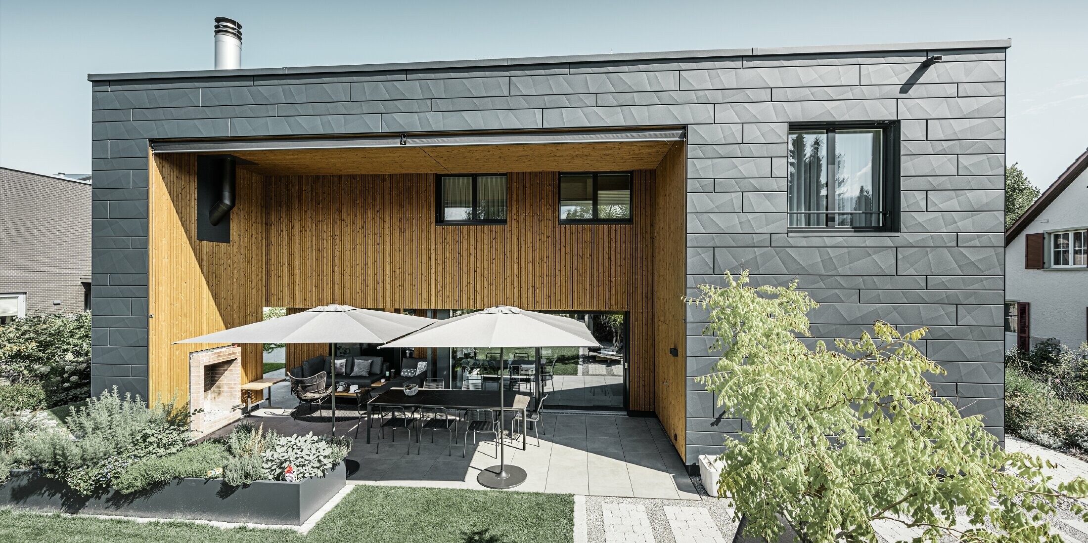 Maison individuelle moderne avec toit plat et façade en Siding.X. La terrasse est joliment agrémentée d’un espace pour s’asseoir.