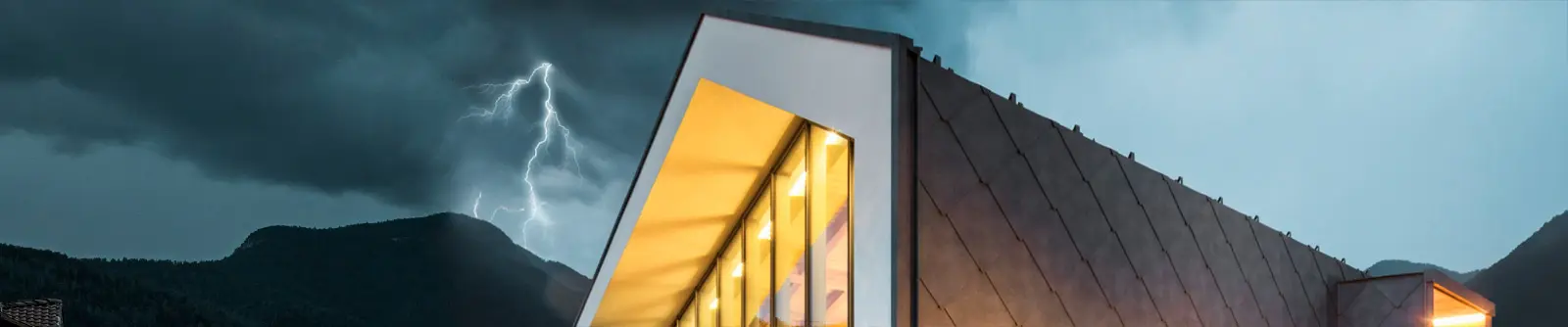 Modernes Gebäude ohne Dachvorsprung und großen Fensterflächen, die Aluminiumfassade ist in Steingrau, im Hintergrund tobt ein Gewitter mit Blitz