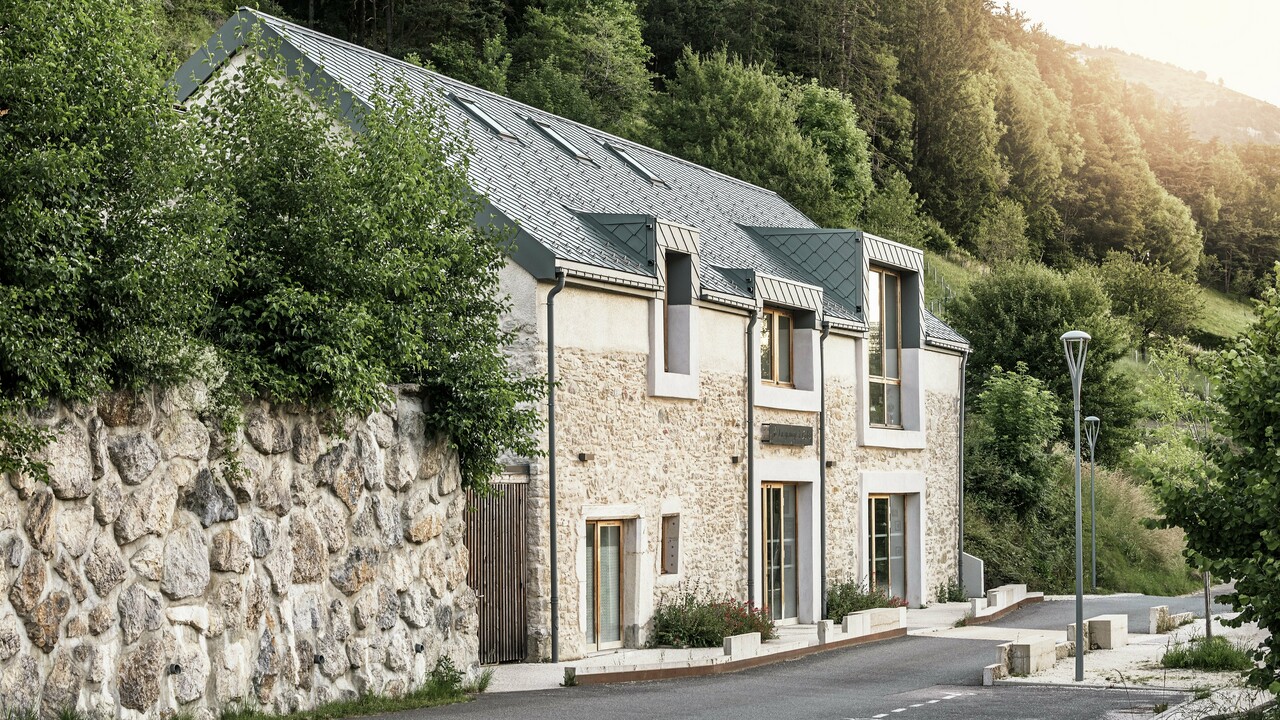 Vue latérale du corps de ferme rénové "La Grange à Gaby" à Engins, France. Le bâtiment combine la maçonnerie en pierre traditionnelle avec un toit moderne en aluminium PREFA composé de losanges de toiture de 29 × 29 cm. La rénovation soignée allie une construction historique à des matériaux durables.
