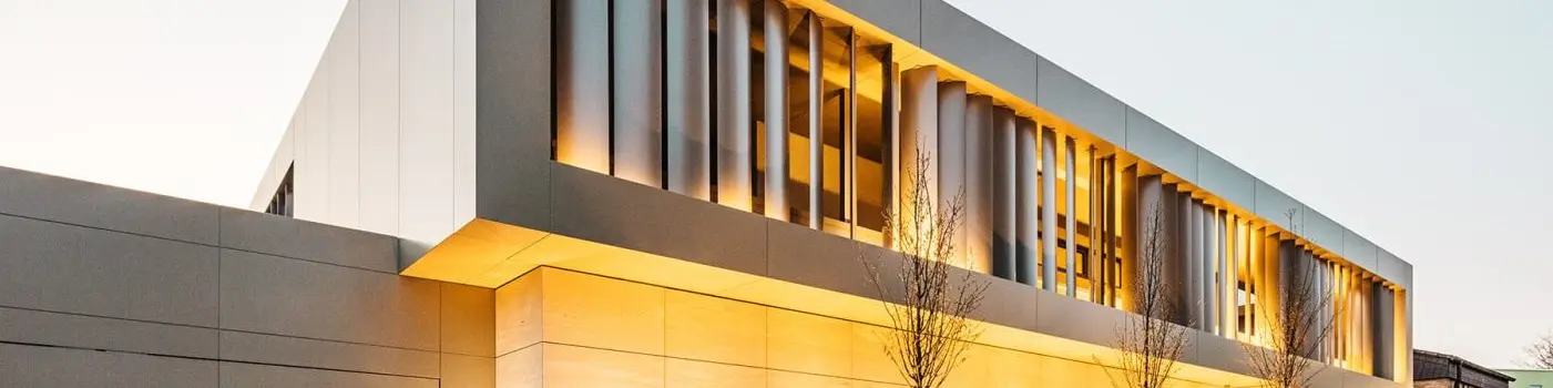 Straßenansicht bei Nacht, links Garage mit bronzefarbener Fassade, unten Steinfassade, oben bronzefarbene Aluminiumfassade mit Lamellen innen