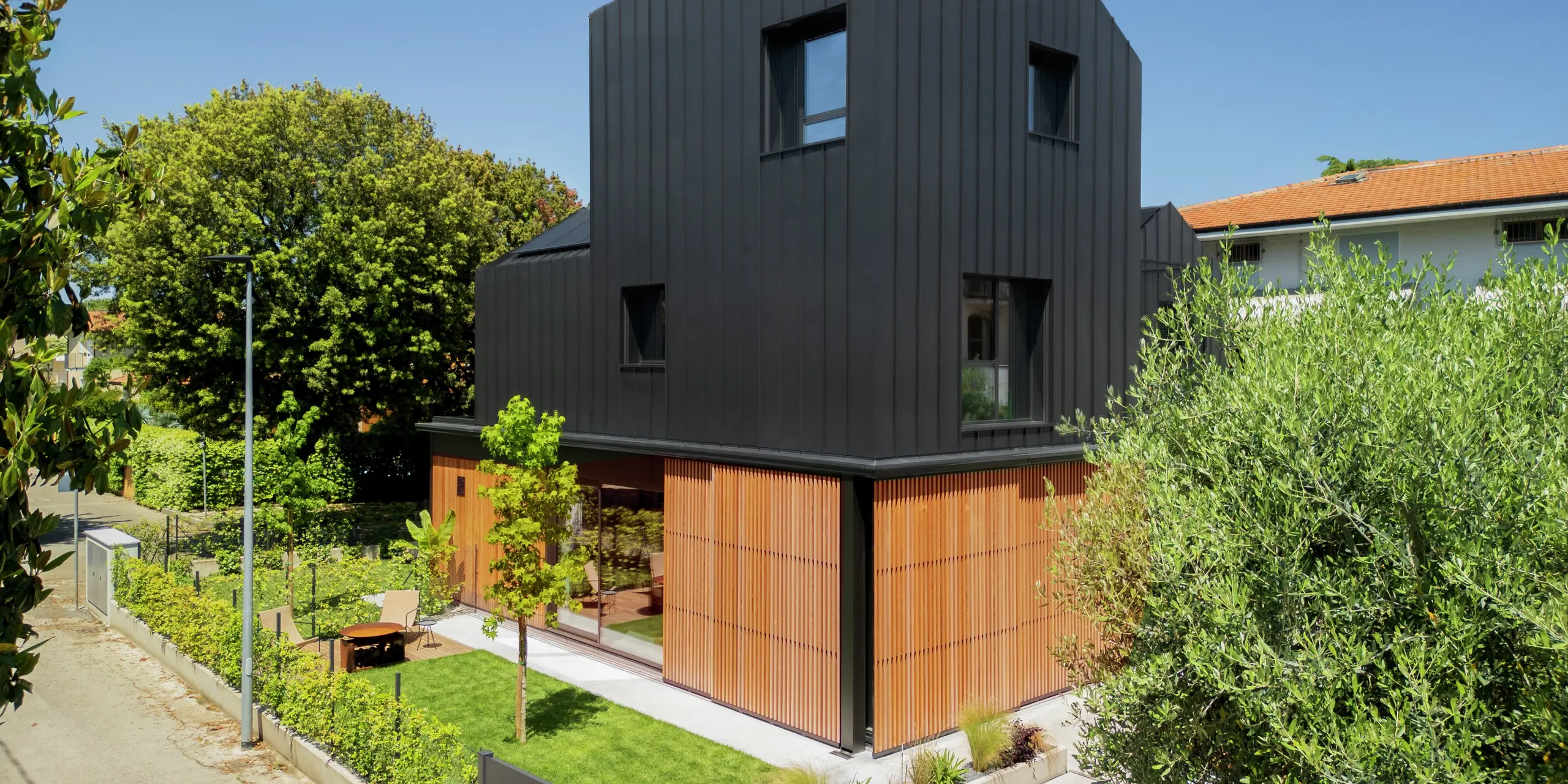 Straßenansicht eines modernen Doppelhauses in Cattolica: elegante Kombination aus Holzlamellen im Erdgeschoss und Aluminiumfassade mit PREFALZ in Schwarzgrau. Die klare Linienführung und die hochwertige PREFA Oberfläche bieten Langlebigkeit, Korrosionsschutz und zeitlose Architektur unter sonnigem Himmel.