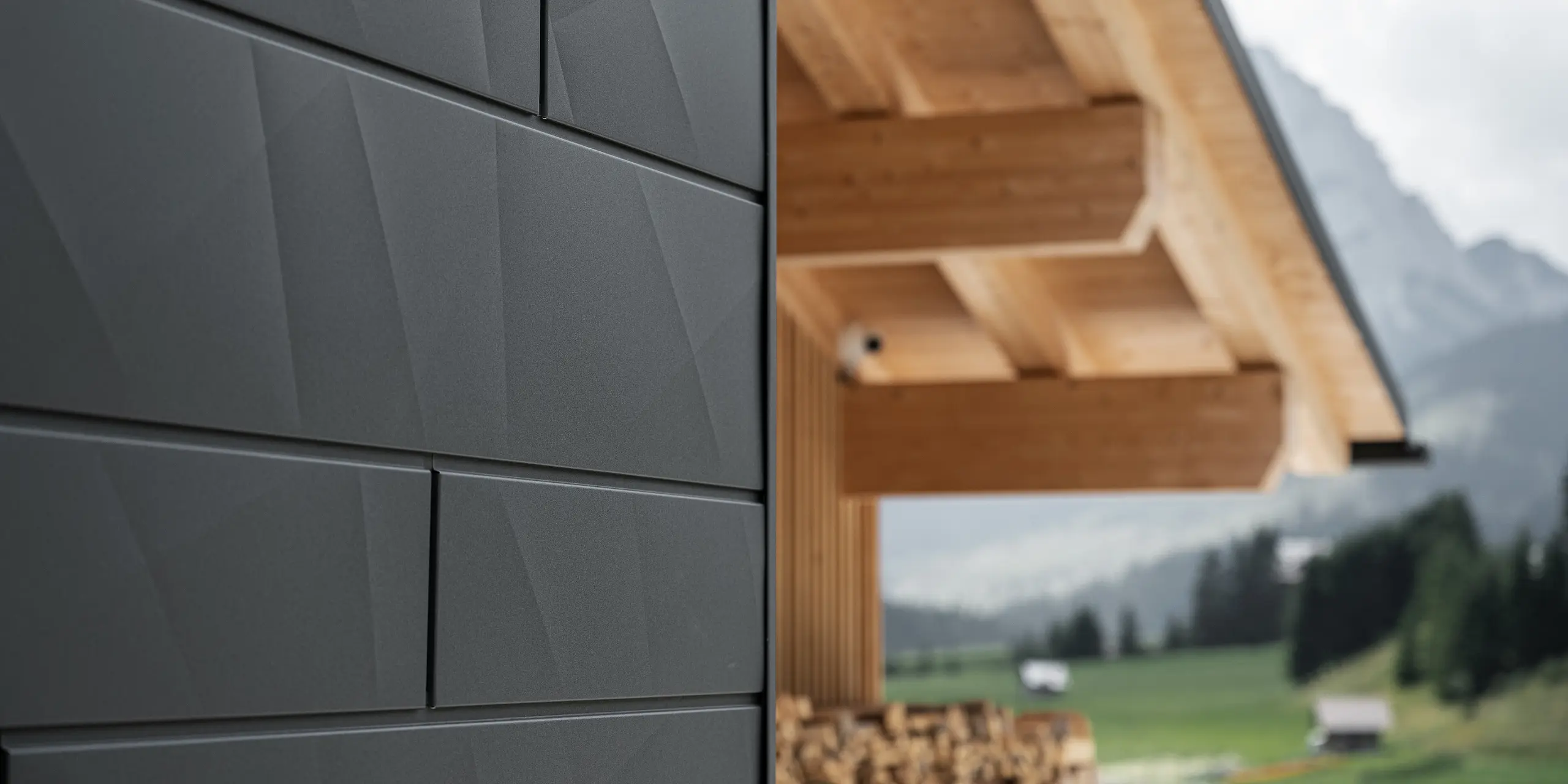 Dettaglio dei pannelli per facciata Doga.X in P.10 nero di PREFA applicati su Casa Hoffe a Sappada (UD). Il sistema di rivestimento in alluminio presenta una posa dal design moderno e astratto, creando un netto contrasto architettonico con le strutture in legno a vista della copertura. La facciata si inserisce armoniosamente nel contesto alpino, coniugando prestazioni tecniche, durabilità e qualità architettonica.