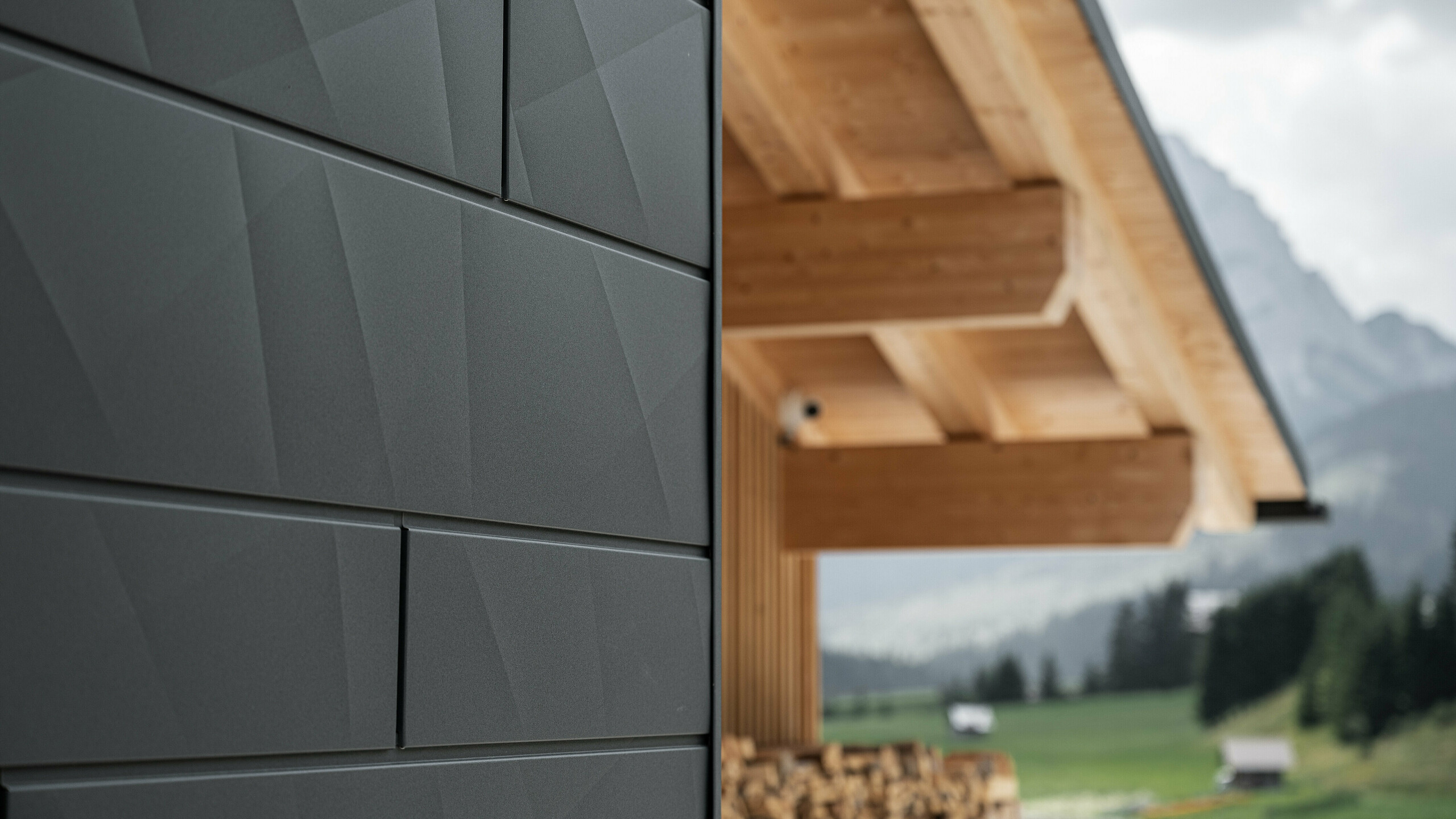 Gros plan sur les panneaux de façade Siding.X en P.10 noir de PREFA sur la Casa Hoffe à Sappada, en Italie. Le système de façade en aluminium est habilement intégré dans un design moderne et abstrait qui contraste agréablement avec les éléments en bois apparents de la charpente de toit. Cette solution de façade innovante s'intègre parfaitement dans l'environnement alpin et allie fonctionnalité, durabilité et esthétique architecturale.