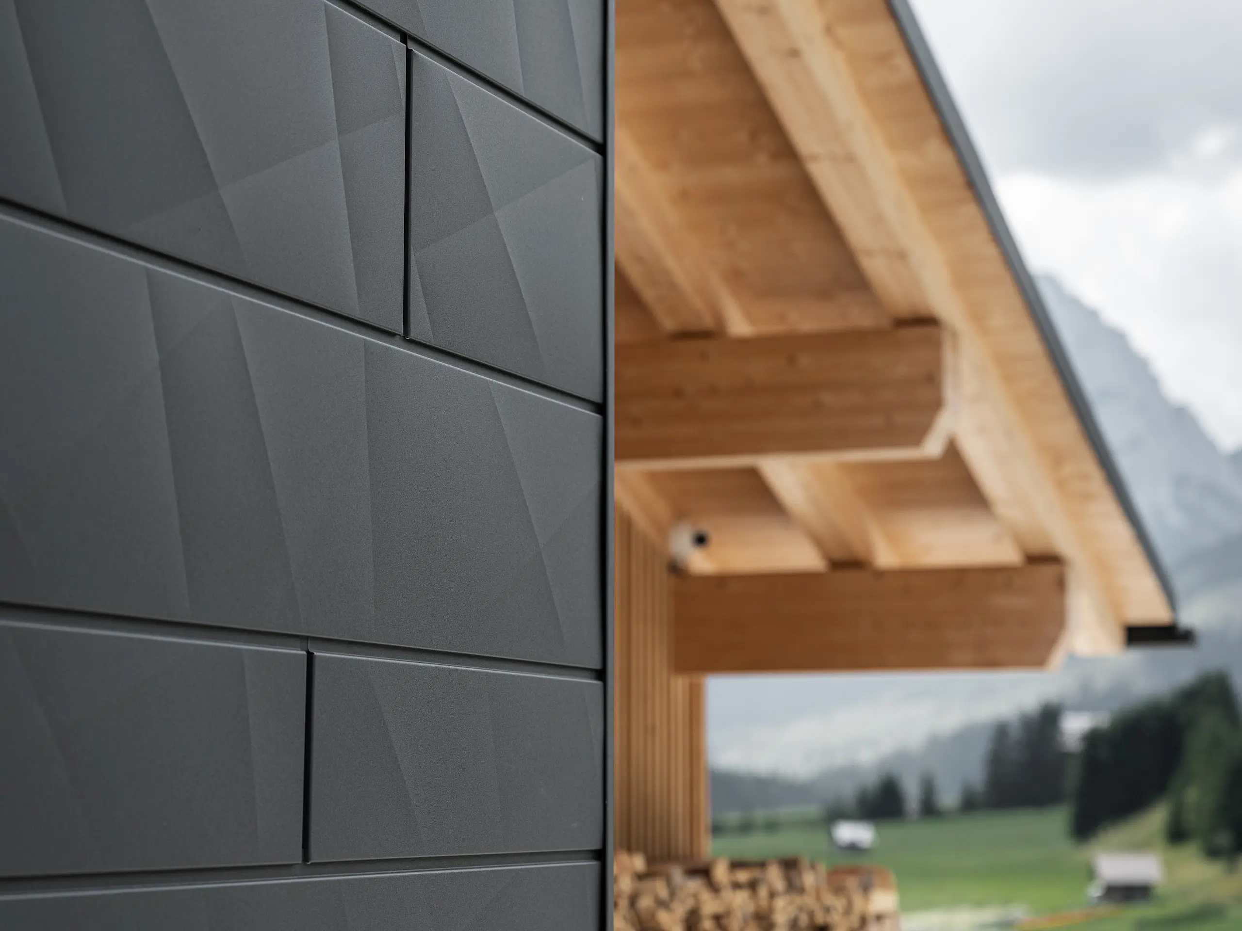 Dettaglio dei pannelli per facciata Doga.X in P.10 nero di PREFA applicati su Casa Hoffe a Sappada (UD). Il sistema di rivestimento in alluminio presenta una posa dal design moderno e astratto, creando un netto contrasto architettonico con le strutture in legno a vista della copertura. La facciata si inserisce armoniosamente nel contesto alpino, coniugando prestazioni tecniche, durabilità e qualità architettonica.