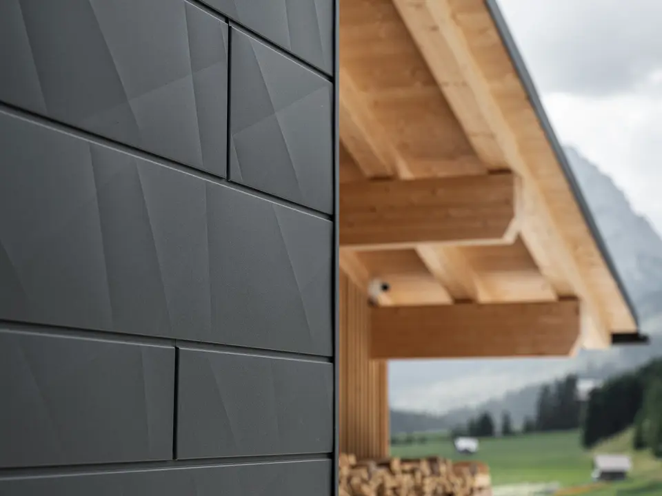 Primo piano dei pannelli di rivestimento Doga.X in P.10 nero di PREFA sulla Casa Hoffe a Sappada, Italia. Il sistema di facciata in alluminio è sapientemente integrato in un design moderno e astratto, che crea un piacevole contrasto con gli elementi in legno a vista della capriata. L'innovativa soluzione per facciate si integra perfettamente nell'ambiente alpino e rappresenta il connubio tra funzionalità, resistenza e cultura edilizia estetica.