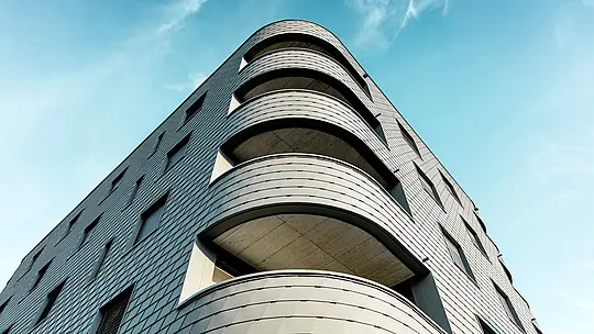 Dynamische Untersicht eines modernen Wohn- und Geschäftshauses mit markant gerundeter Ecke und fließenden Balkonbändern. Die schuppenartige Fassade aus PREFA Wandschindeln in P.10 Hellgrau legt sich wie eine präzise verarbeitete Aluminiumhaut über das Gebäude und betont die weichen Radien. Die homogene Oberfläche unterstreicht die klare Linienführung, während das leichte, witterungsbeständige PREFA Aluminium selbst bei starken Rundungen höchste Formstabilität, Langlebigkeit und Wartungsarmut garantiert – ein architektonischer Eyecatcher vor strahlend blauem Himmel.