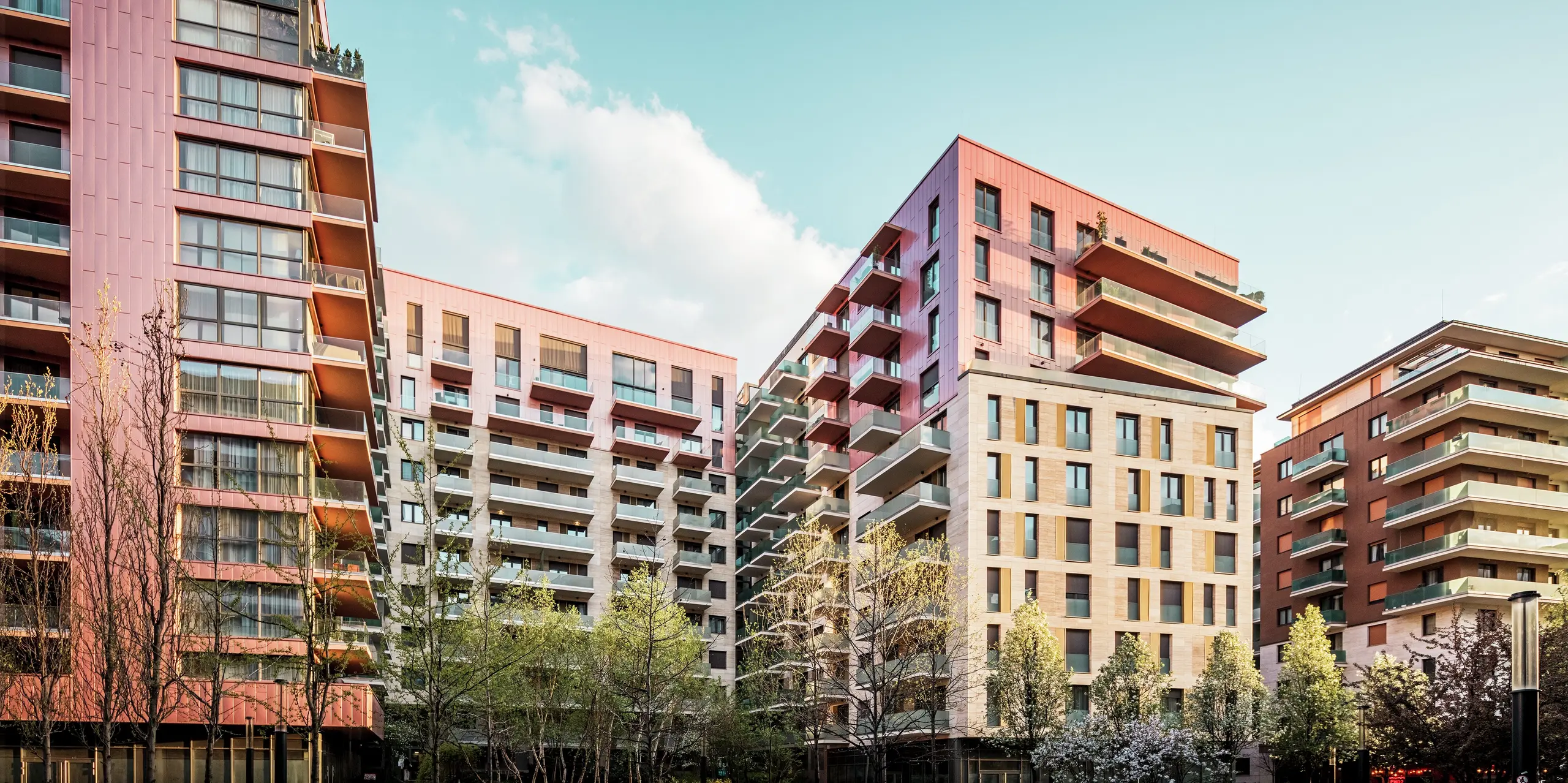 Gesamtansicht des Grand Corvin Wohnprojekts im revitalisierten Corvin-Viertel von Budapest. Die auffällige PREFA Siding Fassadenverkleidung in der Sonderfarbe Lachsmetallic setzt starke visuelle Akzente und betont die oberen Etagen. In Kombination mit Naturstein und Glas entsteht eine hochwertige, moderne Architektur. Die langlebigen Aluminiumpaneele sind pflegeleicht, witterungsbeständig und fügen sich harmonisch in das urbane Umfeld ein. Das Bild wurde während der Golden Hour aufgenommen, weshalb angenehmes, warmes Licht die Szene einrahmt.