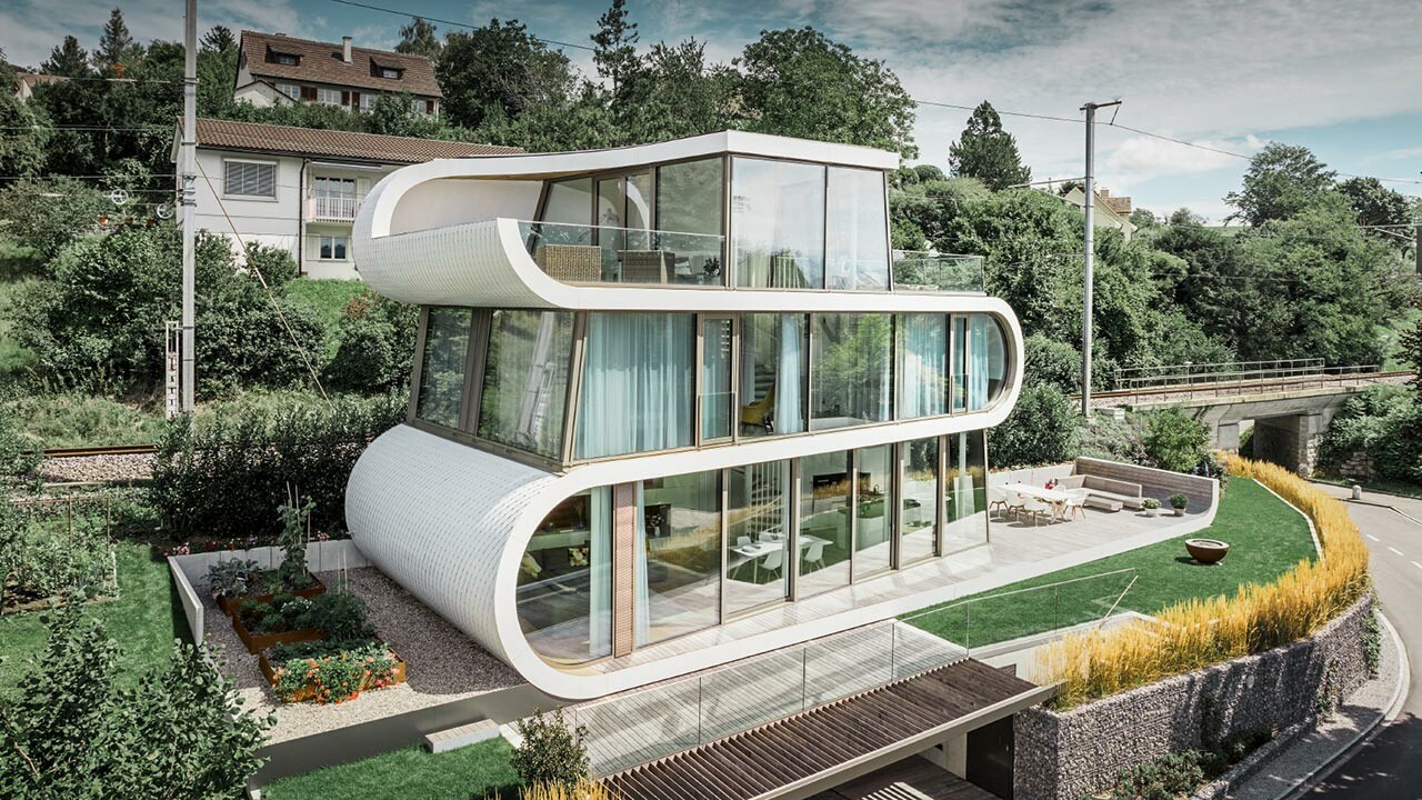 Aufnahme des modernen Flexhouses am Zürichsee. Mit seiner außergewöhnlichen Gebäudeform fällt das Flexhouse immer auf. Die Geschoßtrennungen ziehen sich als weißes Band geschwungen durch das Gebäude. Die runden Fassadenelemente wurden mit den Alu-Schindeln von PREFA in Reinweiß verkleidet. 