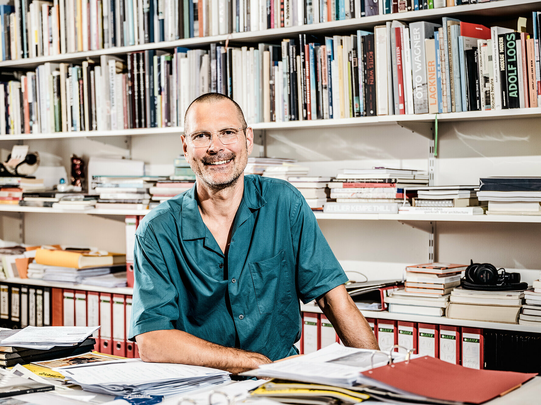 Portrait von Architekt Eckehart Loidolt in seinem Bürozimmer, umgeben von Arbeitsmaterial und Büchern auf dem Regal im Hintergrund.