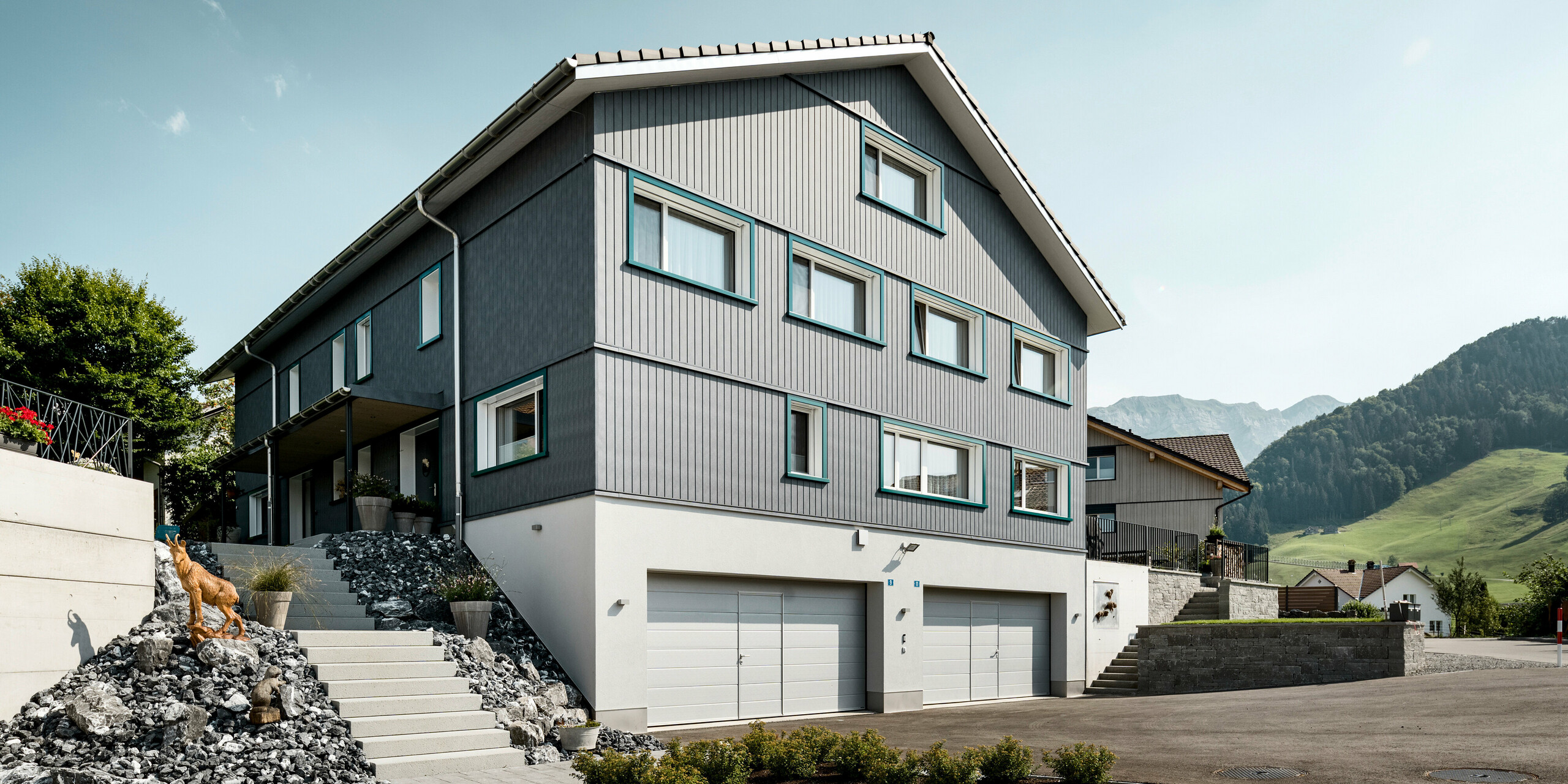 Vorderansicht eines Mehrfamilienhauses in Weissbad, Schweiz, mit einer markanten PREFA Siding Fassade in P.10 Steingrau. Die vertikal verlegten Aluminiumsidings heben die straffe, moderne Form des Gebäudes hervor und kontrastieren ansprechend mit den lebendigen türkisen Fensterrahmen. Die Fassade fügt sich harmonisch in die alpine Landschaft ein und bietet mit ihrer Beständigkeit und ihrem stilvollen Design einen dauerhaften Wohnwert.