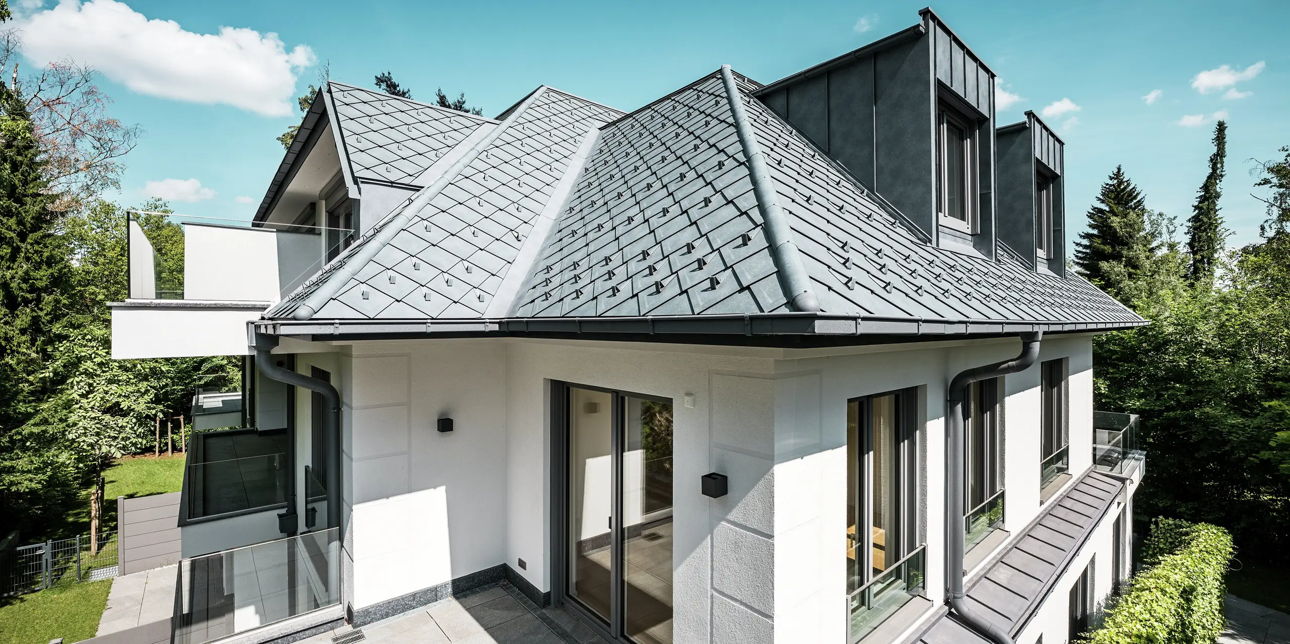 Maison multifamiliale moderne à l'architecture de toiture remarquable, réalisée en PREFA P.10 Gris Pierre : la surface du toit est recouverte de losanges PREFA 29 x 29 cm de haute qualité, créant un aspect texturé et élégant. Des lucarnes plates stylées en PREFALZ s'intègrent harmonieusement au design. Des arrêts de neige intégrés et un système d'évacuation des eaux pluviales PREFA esthétique, avec gouttières et descentes, soulignent la sophistication technique. La toiture en aluminium durable résiste à toutes les conditions climatiques et allie esthétique et fonctionnalité – idéale pour la construction résidentielle contemporaine.
