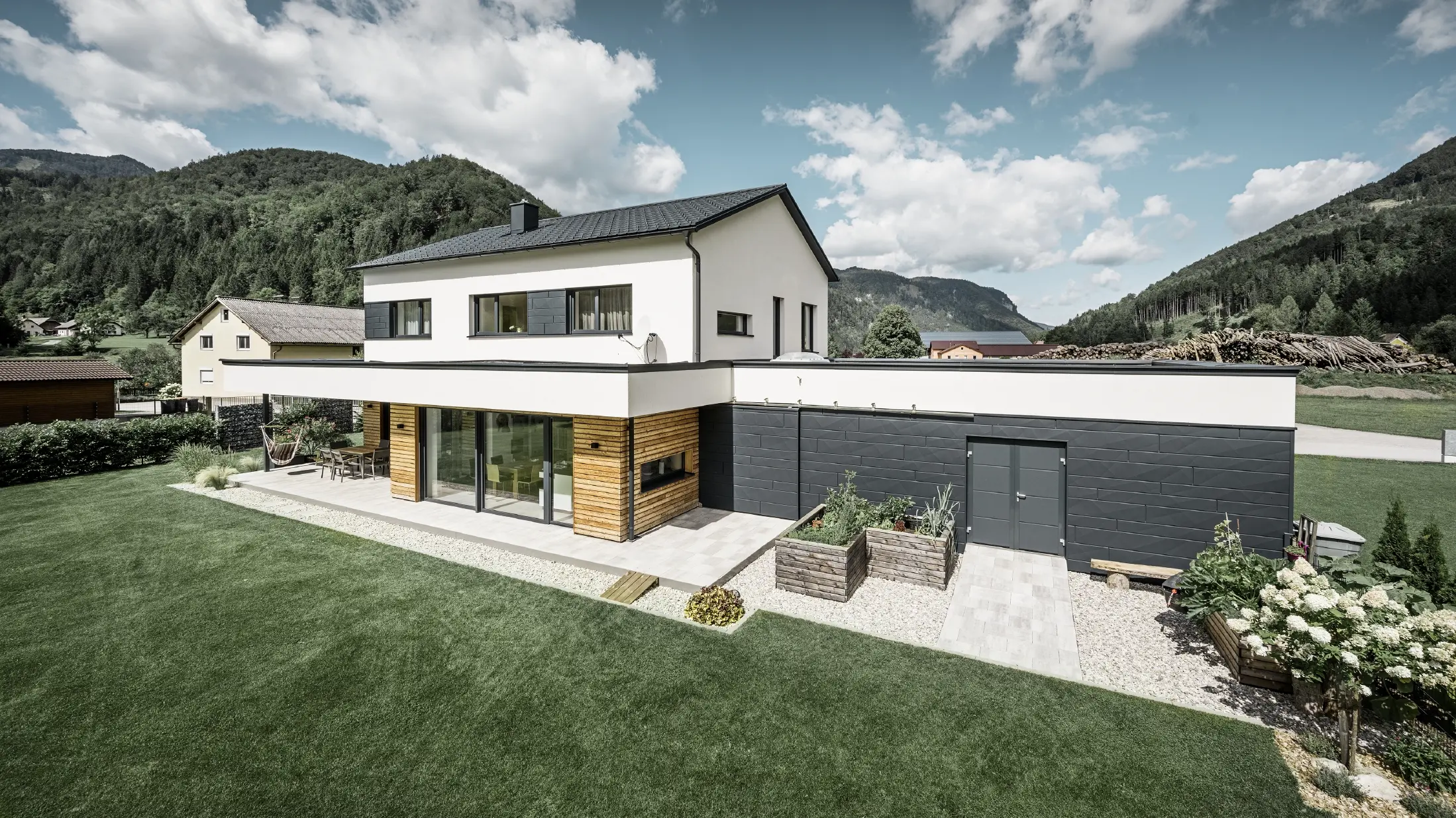 Das Bild zeigt ein modernes Einfamilienhaus in einer idyllischen Landschaft mit einer klaren Sicht auf bewaldete Hügel im Hintergrund. Das Haus zeichnet sich durch eine Kombination aus weißen Putzwänden und einer markanten Fassadenverkleidung mit PREFA Siding.X in P.10 Anthrazit aus, die für einen modernen und langlebigen Look sorgt. Die dunkle Färbung der Fassadenelemente bildet einen ansprechenden Kontrast zum hellen Außendesign und zur natürlichen Umgebung. Die Anordnung der Siding.X Fassadenpaneele ist präzise und verleiht der Immobilie einen strukturierten und hochwertigen Charakter, der typisch für PREFA Produkte ist. Das Dach des Gebäudes wurde mit der bewährten PREFA Dachplatte eingedeckt. Die Dachentwässerung erfolgt durch das Ablaufrohr und die Hängerinne von PREFA.