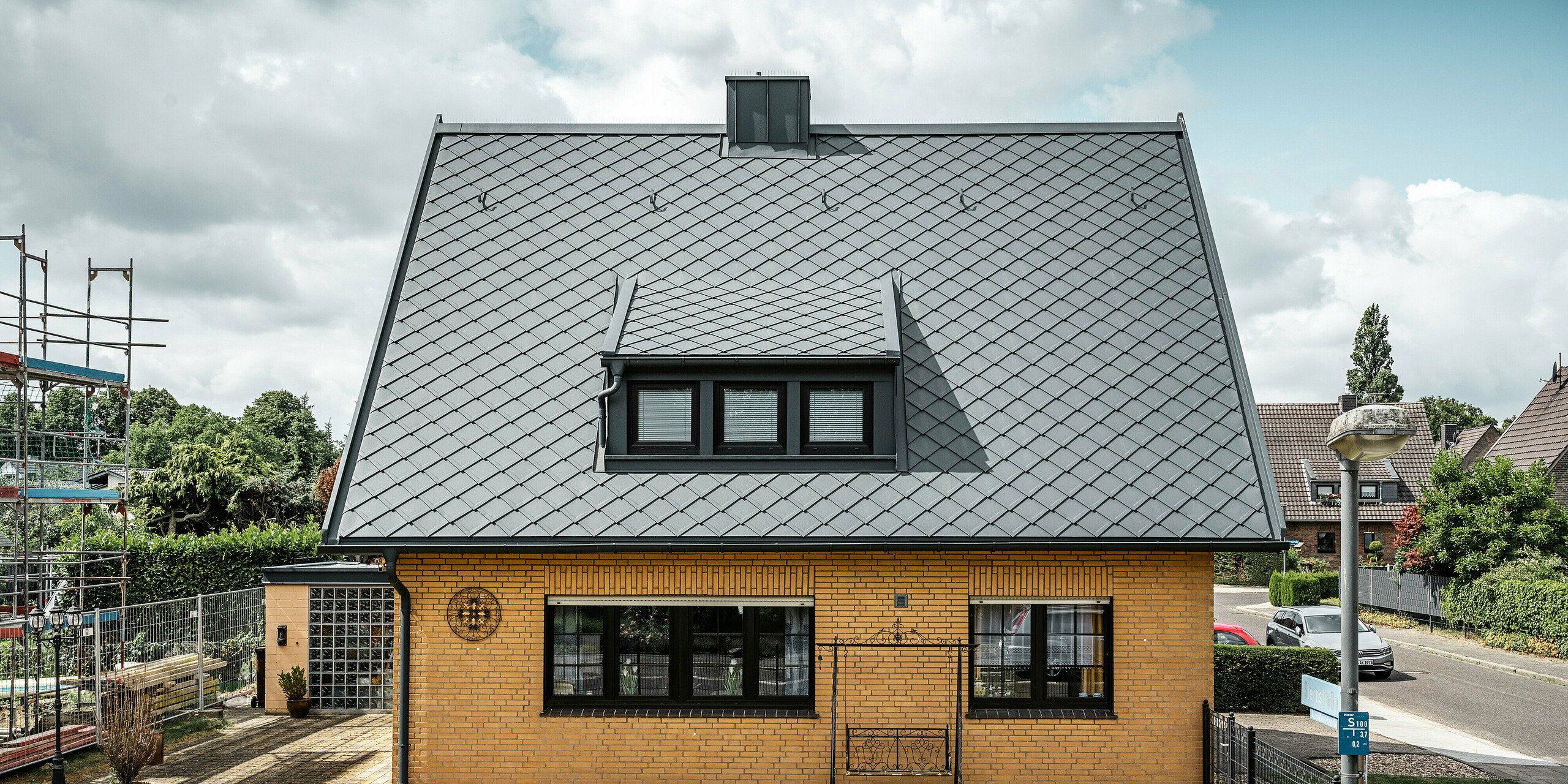 Seitenansicht eines Einfamilienhauses in Tönisvorst mit einem anthrazitfarbenen PREFA Dach aus Dachrauten 29×29. Die Kombination des traditionellen gelben Backsteins mit den modernen Dachelementen und Zubehör aus Aluminium ergibt ein harmonisches Gesamtbild. Schleppgauben und Kamin, ebenfalls in Anthrazit gehalten, vervollständigen das ästhetische und funktionale Dach in einer kleinstädtischen Umgebung.