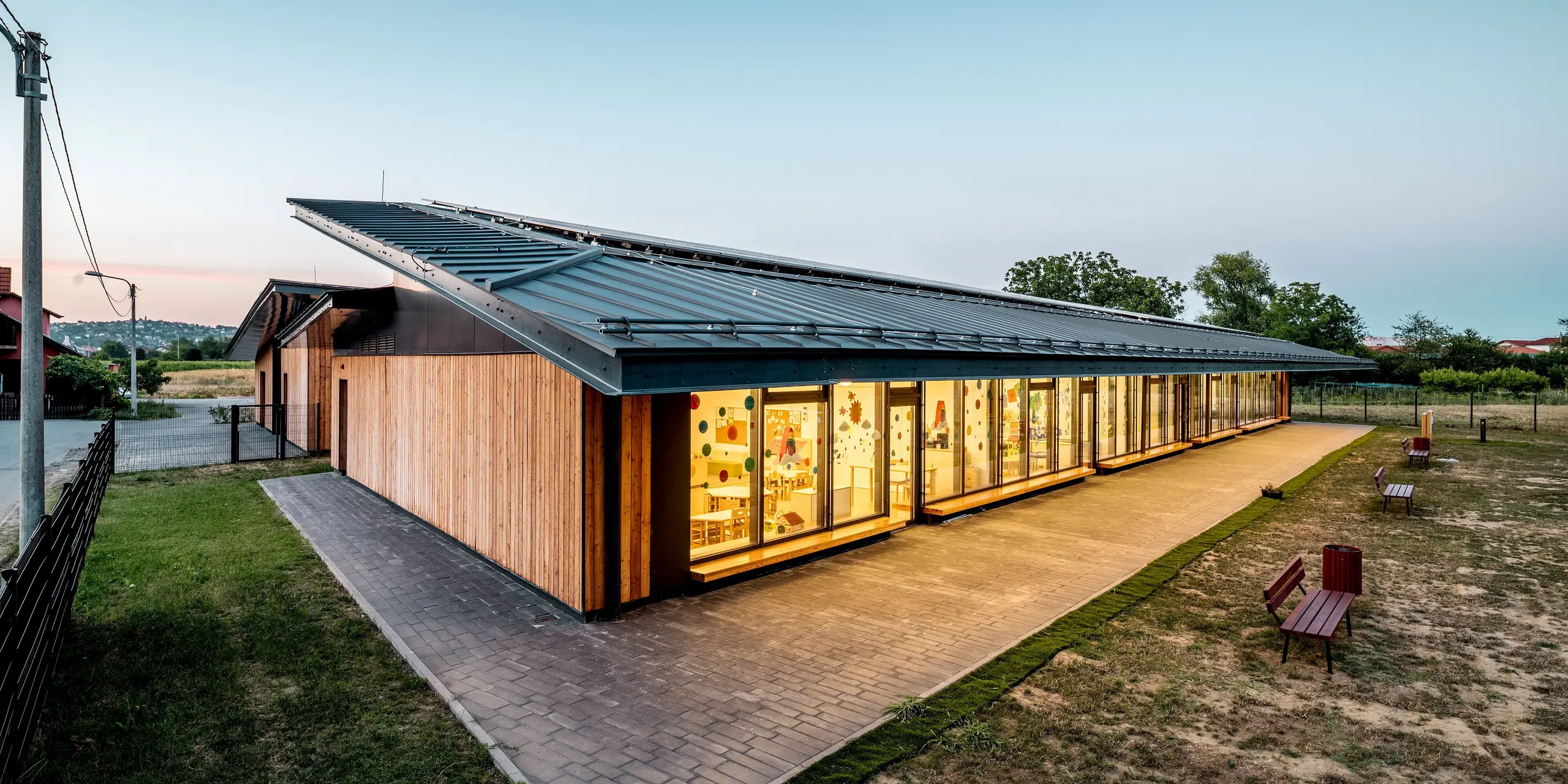 Abendliche Außenansicht des Kindergartens Svarozic mit elegantem PREFALZ Aluminiumdach in P.10 Anthrazit. Die großzügige Dachfläche schützt die helle Glasfassade und die natürliche Holzverkleidung, während warme Innenbeleuchtung eine einladende Atmosphäre schafft. Das PREFA Dach überzeugt durch klare Linien, hohe Widerstandsfähigkeit und langlebigen Schutz für die moderne Architektur.