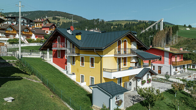 Einfamilienhaus in Gallio in traditioneller Alpenarchitektur mit leuchtend gelb-roter Putzfassade und Natursteinelementen. Das Dach aus PREFA Dachplatten R.16 in P.10 Dunkelgrau bietet höchste Witterungsbeständigkeit und harmoniert perfekt mit der Umgebung. Die Dachentwässerung mit PREFA Hängerinne und Ablaufrohr - ebenfalls in P.10 Dunkelgrau - sorgt für zuverlässigen Schutz. Umgeben von grünen Wiesen und mit Blick auf die Berge vereint das Haus Ästhetik, Langlebigkeit und Funktionalität. Im Hintergrund ist die bekannte Skisprungschanze der Region zu erkennen.