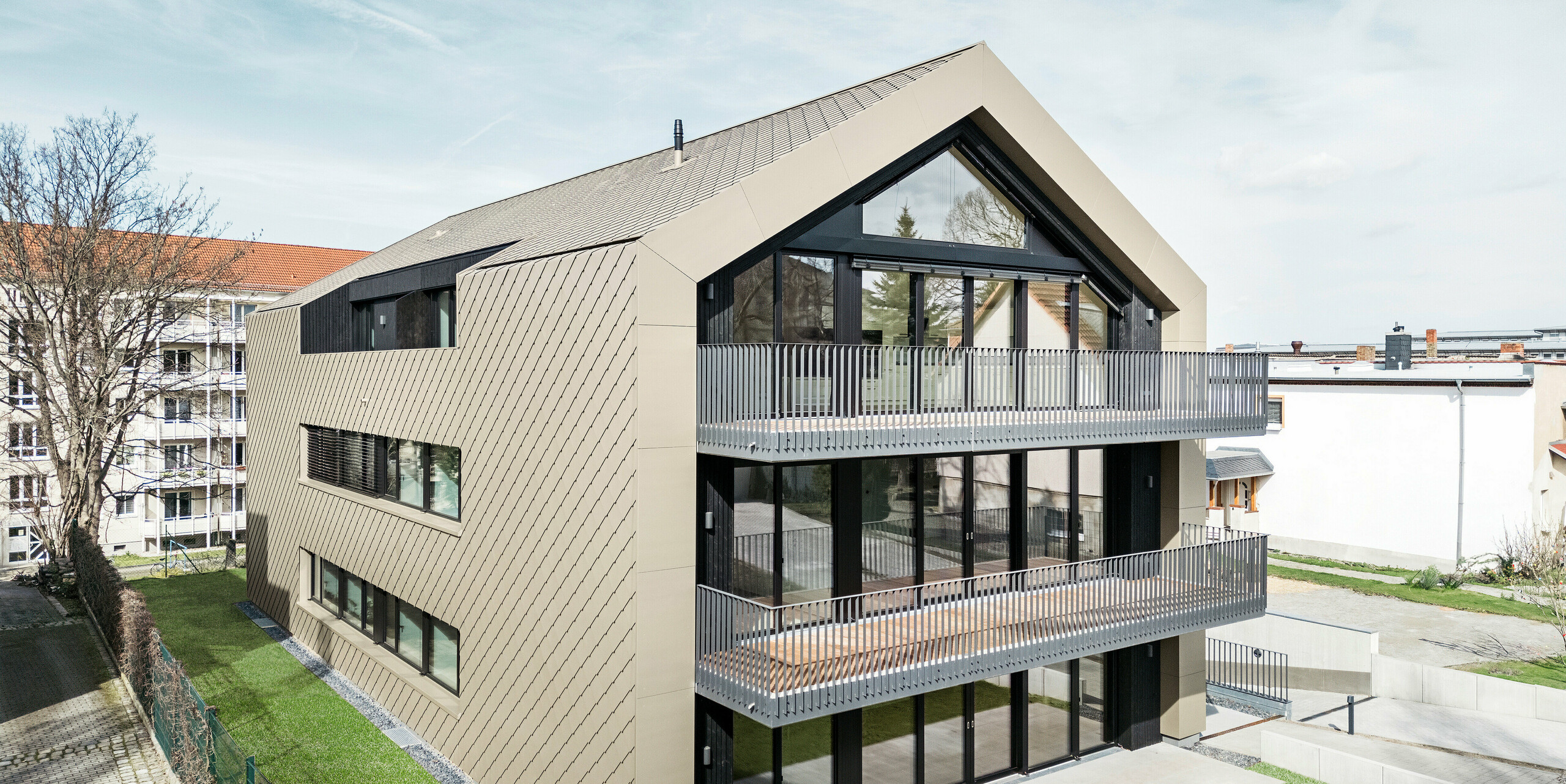 Vue sur le jardin d'un immeuble d'appartements moderne sur la Chemnitzer Straße à Dresde. Le jardin bien entretenu est entouré d'une architecture impressionnante. La façade et le toit du bâtiment sont revêtus du losange en aluminium durable PREFA 29 × 29 en bronze, qui confère à la structure un aspect élégant et contemporain. La photo montre les balcons spacieux, les grandes façades vitrées et l'intégration harmonieuse du bâtiment dans l'environnement.