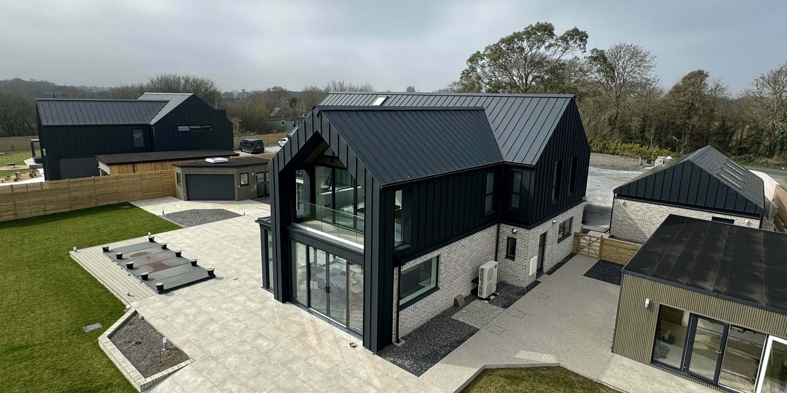 Vue d'une maison individuelle moderne avec une toiture et une façade PREFALZ P.10 gris sombre, associées à un parement en pierre claire au rez-de-chaussée. Les larges baies vitrées, qui laissent entrer une abondante lumière naturelle, sont particulièrement remarquables. Au premier plan, une terrasse spacieuse aux lignes épurées, tandis qu'à l'arrière-plan se dessinent d'autres bâtiments du complexe Bullenmead. Les surfaces en aluminium sont durables, résistantes à la corrosion et parfaitement adaptées au climat côtier. Le contraste entre la toiture sombre, le verre et la pierre naturelle souligne l'élégance de l'architecture.