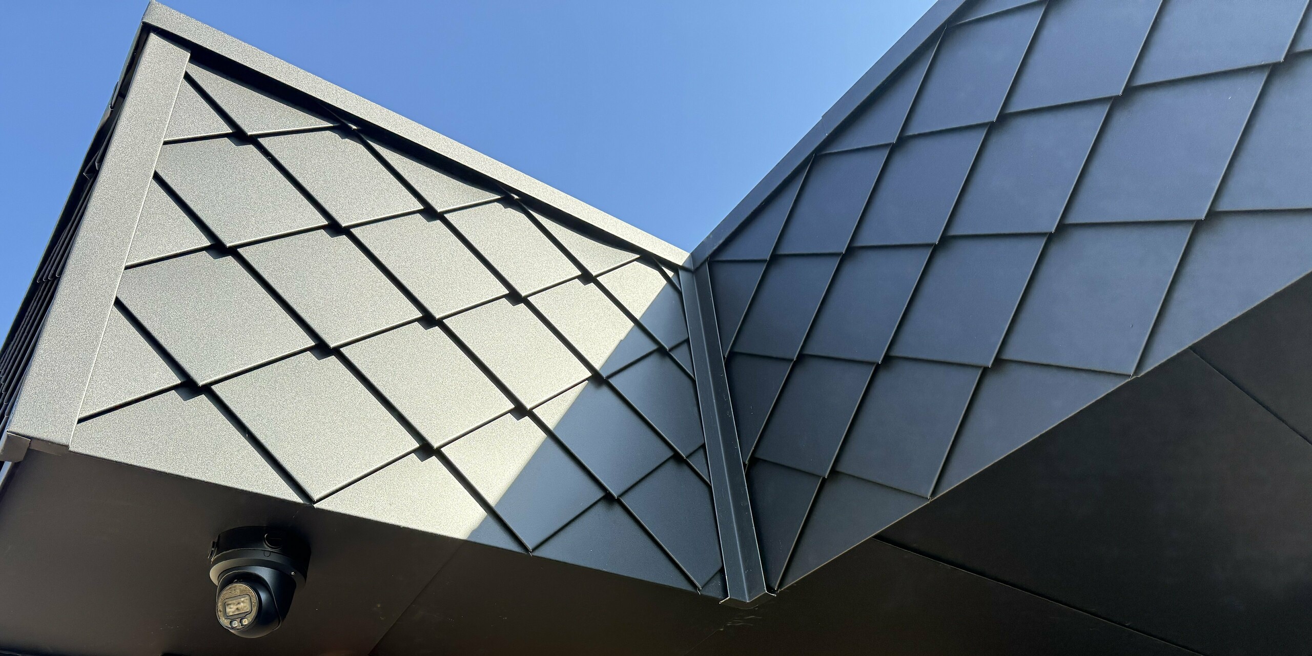 Vue détaillée d'un angle de bâtiment à la façade remarquable : le petit format de losange de façade PREFA 20 × 20 en P.10 anthracite apporte une touche d'élégance et souligne le dynamisme des lignes. Contrastant avec la chaleur de la façade en briques, il crée un mélange de matériaux saisissant, alliant design moderne et qualité supérieure. Idéal pour une architecture durable au caractère affirmé.