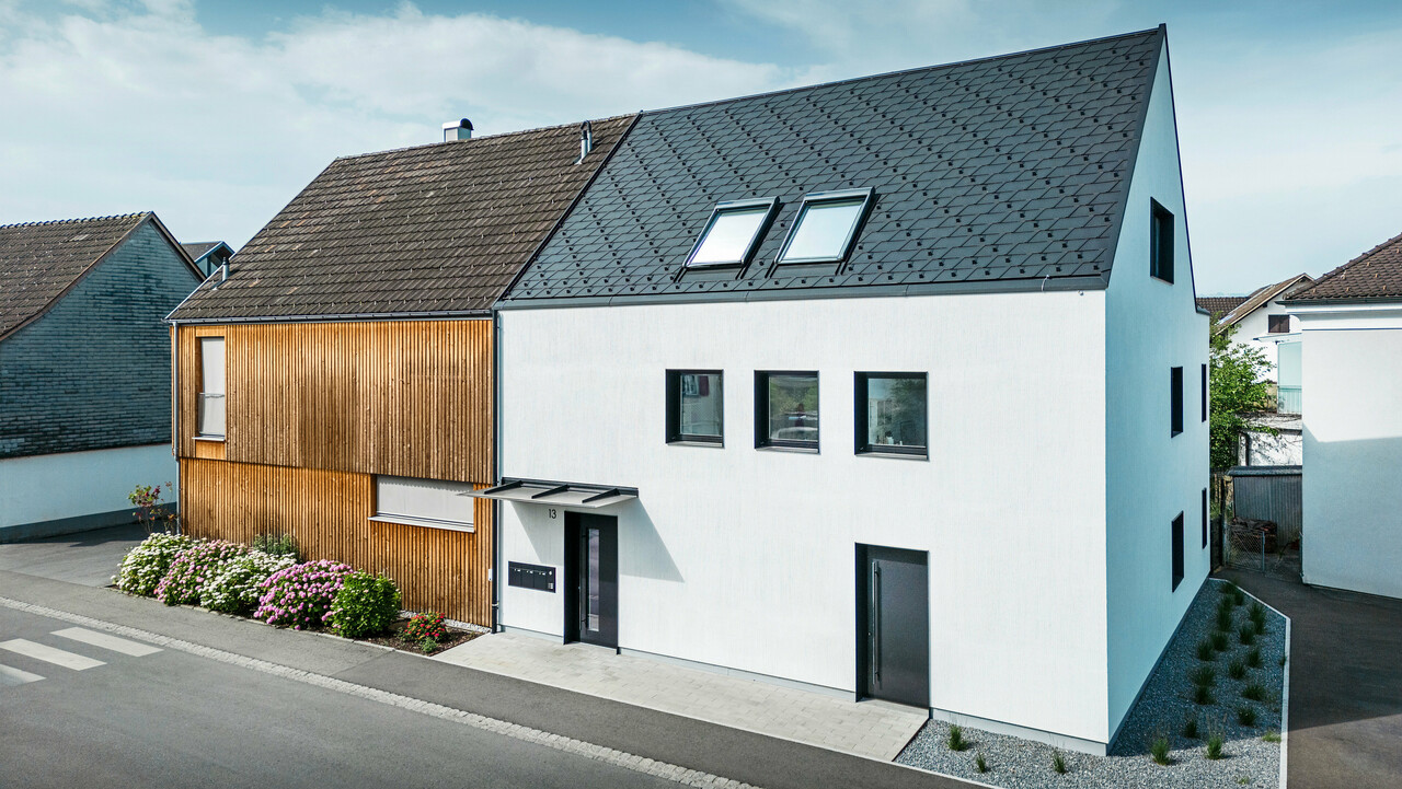 Vue aérienne d'un immeuble collectif d'angle avec un toit à deux pans à arêtes vives, couvert de bardeaux PREFA DS19 et de Prefalz en noir. Un côté de la maison est recouvert de lattes de bois verticales, l'autre côté est crépi d'un blanc pur, avec plusieurs fenêtres encastrées dans la façade. Devant la maison se trouve un jardin bien entretenu avec une grande variété de fleurs et d'arbustes. L'image est complétée par un ciel bleu clair en arrière-plan.