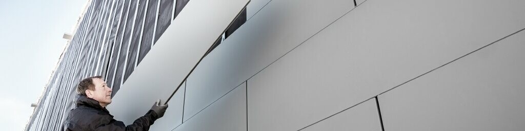  Foto di una facciata parzialmente rivestita con doghe PREFA larghezza 500. L'installatore posa un altro pannello di facciata. La foto è scattata dal basso.