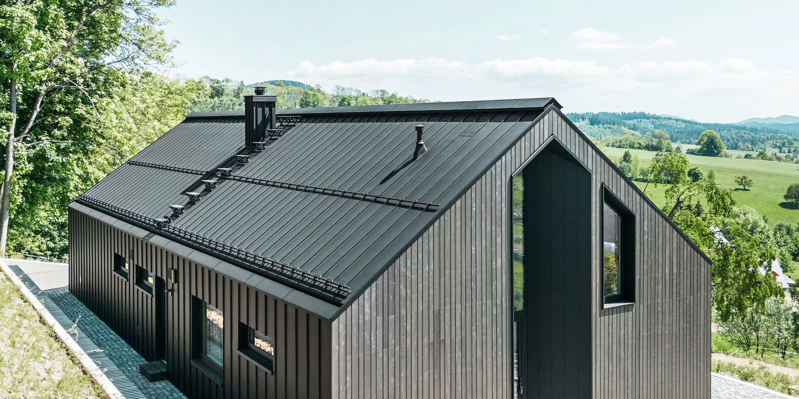 A dark solid-timber house clad in PREFALZ in P. 10 on the roof and the reverse of the building. In front of the house stands a small outbuilding with an aluminium cladding of PREFALZ in P.10 black. In the background, a bright blue sky and woodland.