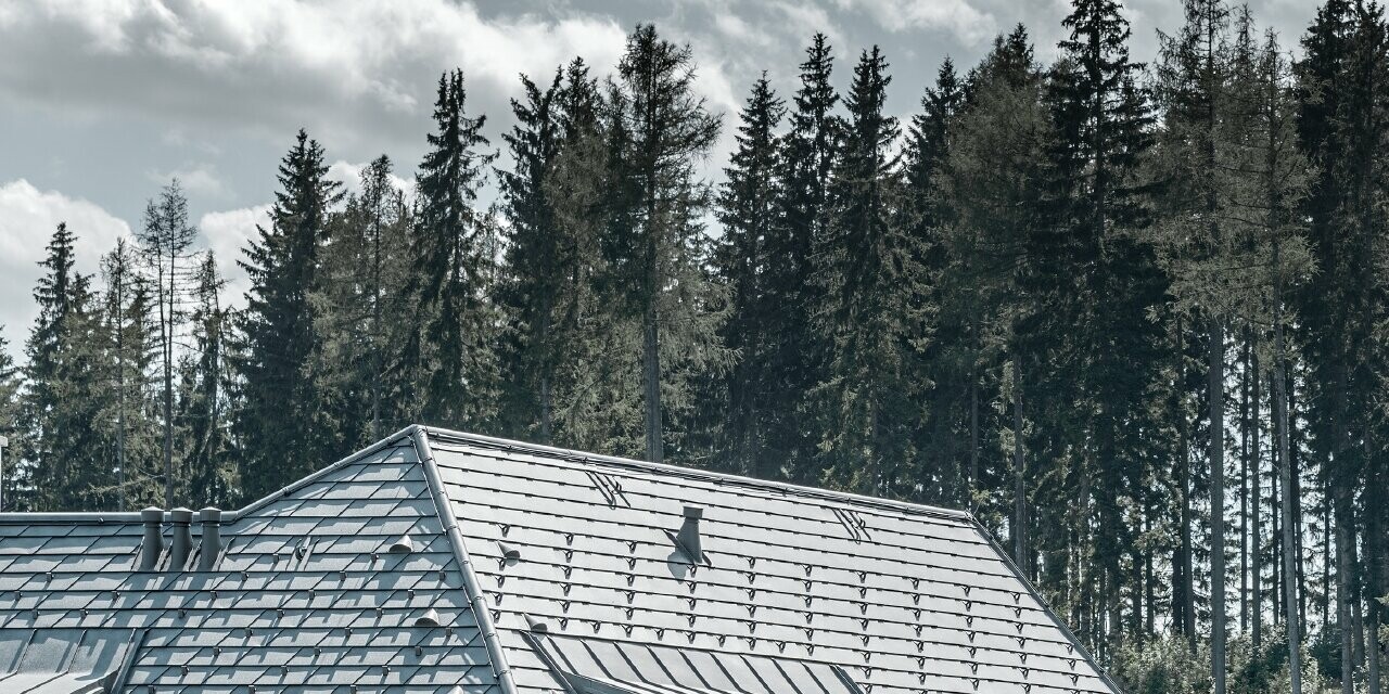 Veduta posteriore della casa unifamiliare in pendenza; il tetto è stato coperto con la scandola in alluminio color grigio pietra, gli abbaini con lamiera con aggraffatura, sempre in grigio pietra. Sullo sfondo si staglia un bosco.