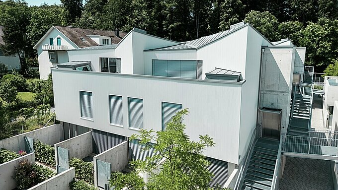 Condominio dall'architettura moderna, dotato di una facciata verticale con profilo a zeta in patina grigio e un tetto in PREFALZ in tonalità abbinata. Le linee pulite dell'edificio si armonizzano con gli spazi esterni integrati, gli elementi in calcestruzzo e le aree giardino strutturate al piano terra. Sullo sfondo, una fitta vegetazione e un edificio residenziale tradizionale sottolineano il contrasto con lo stile architettonico moderno.