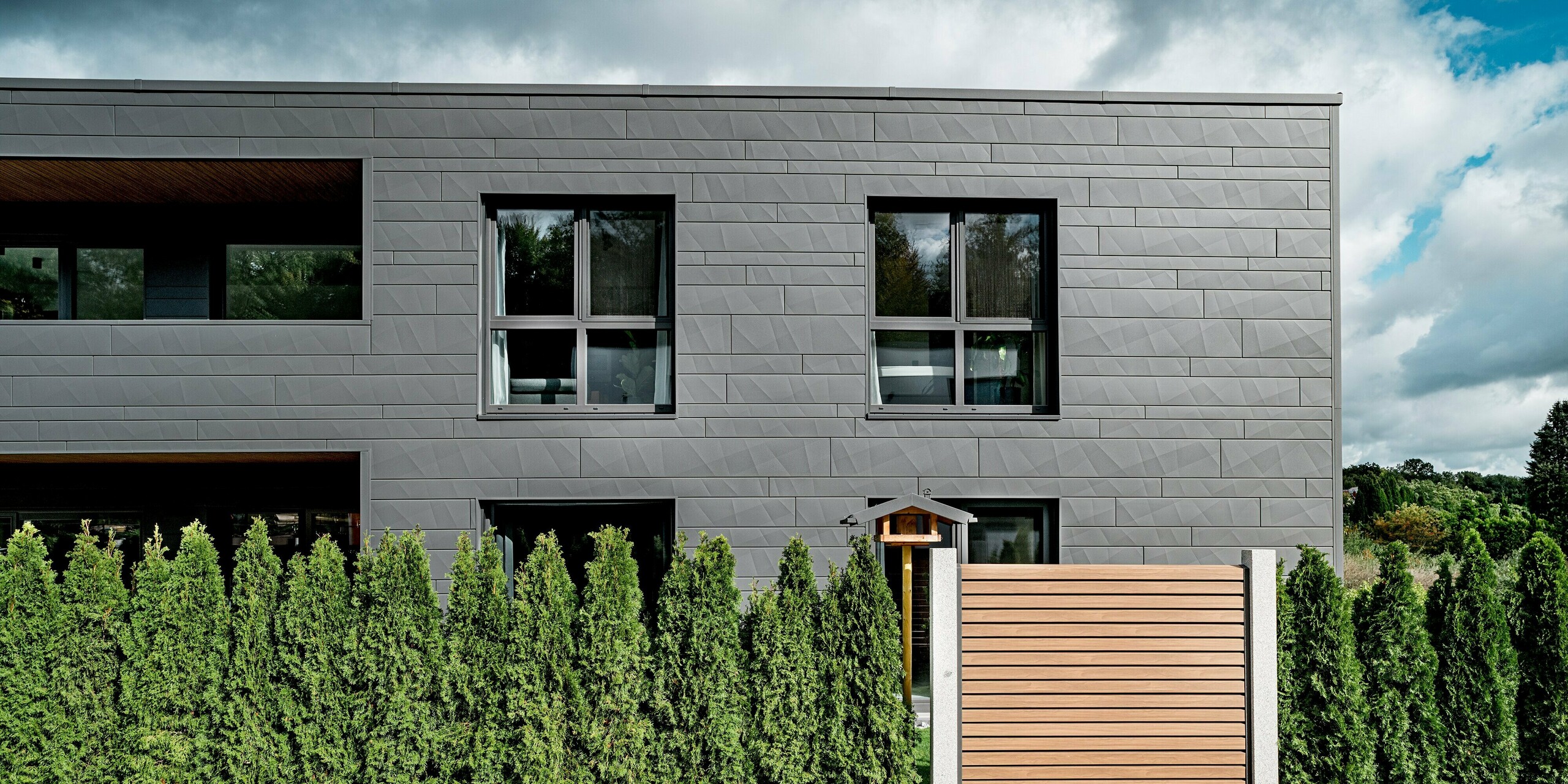 Seitenansicht eines modernen Einfamilienhauses in Heidenheim mit vollständig verkleideter PREFA Siding.X Fassade in P.10 Dunkelgrau. Die horizontal angeordneten Aluminiumpaneele mit unterschiedlichen Breiten und markanten Kantungen erzeugen eine lebendige, strukturierte Oberfläche. Im Vordergrund verläuft eine dichte Reihe immergrüner Thujen, während ein Holzzaun einen warmen Kontrast zur kühlen Fassadenarchitektur setzt. Große Fensteröffnungen spiegeln die Umgebung und betonen die klare Linienführung. Die langlebige PREFA Aluminiumoberfläche sorgt für dauerhaften Schutz und moderne Eleganz.