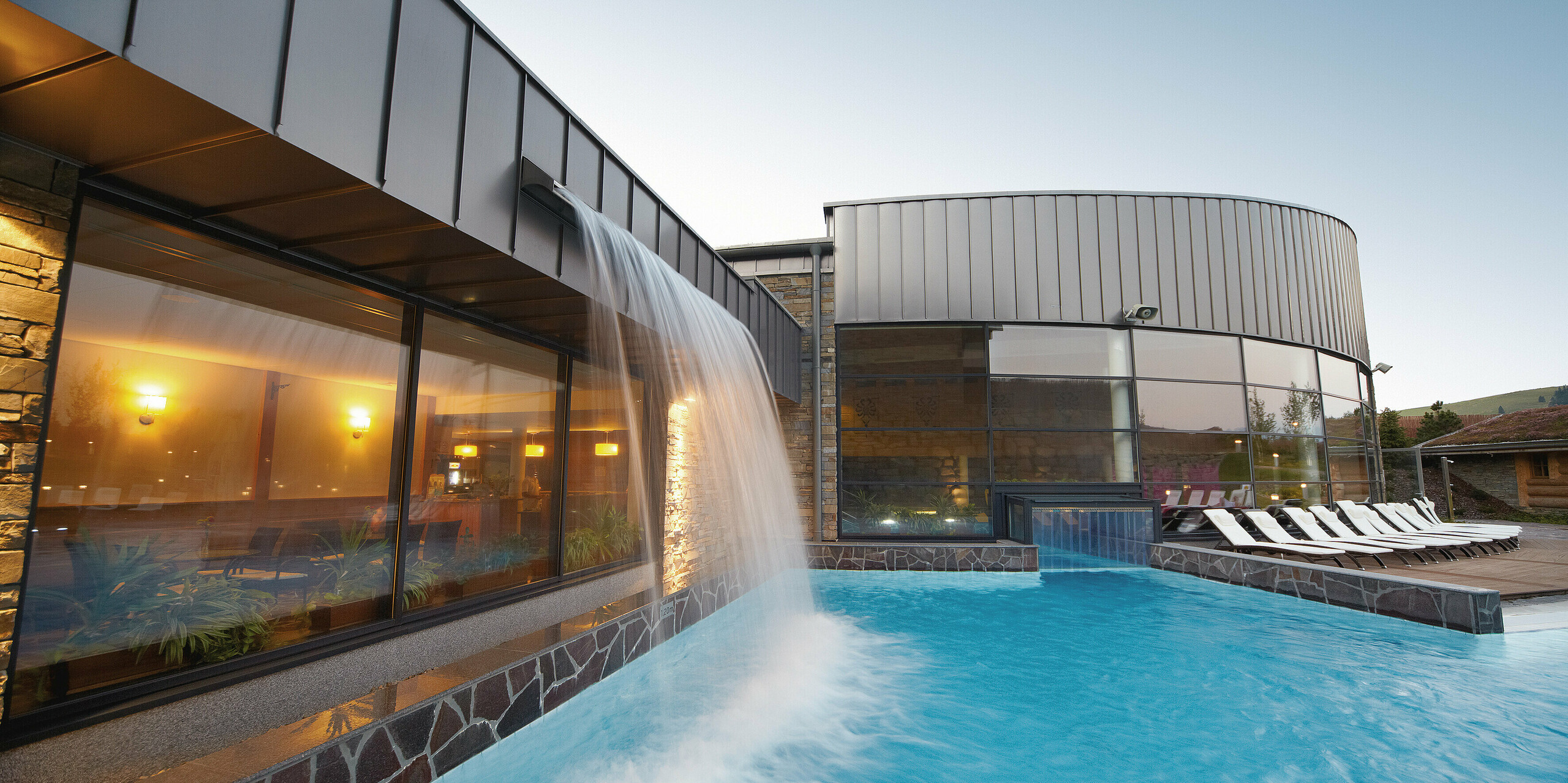 Ein Blick auf den imposanten Wasserfall der Therme Białka zeigt das perfekte Zusammenspiel von Architektur und Natur. Die PREFALZ-Fassade in der Sonderfarbe Delphin ergänzt die organischen Elemente und bildet einen eleganten Kontrast zur Steinmauer und den sanften Wasserbewegungen im Poolbereich.