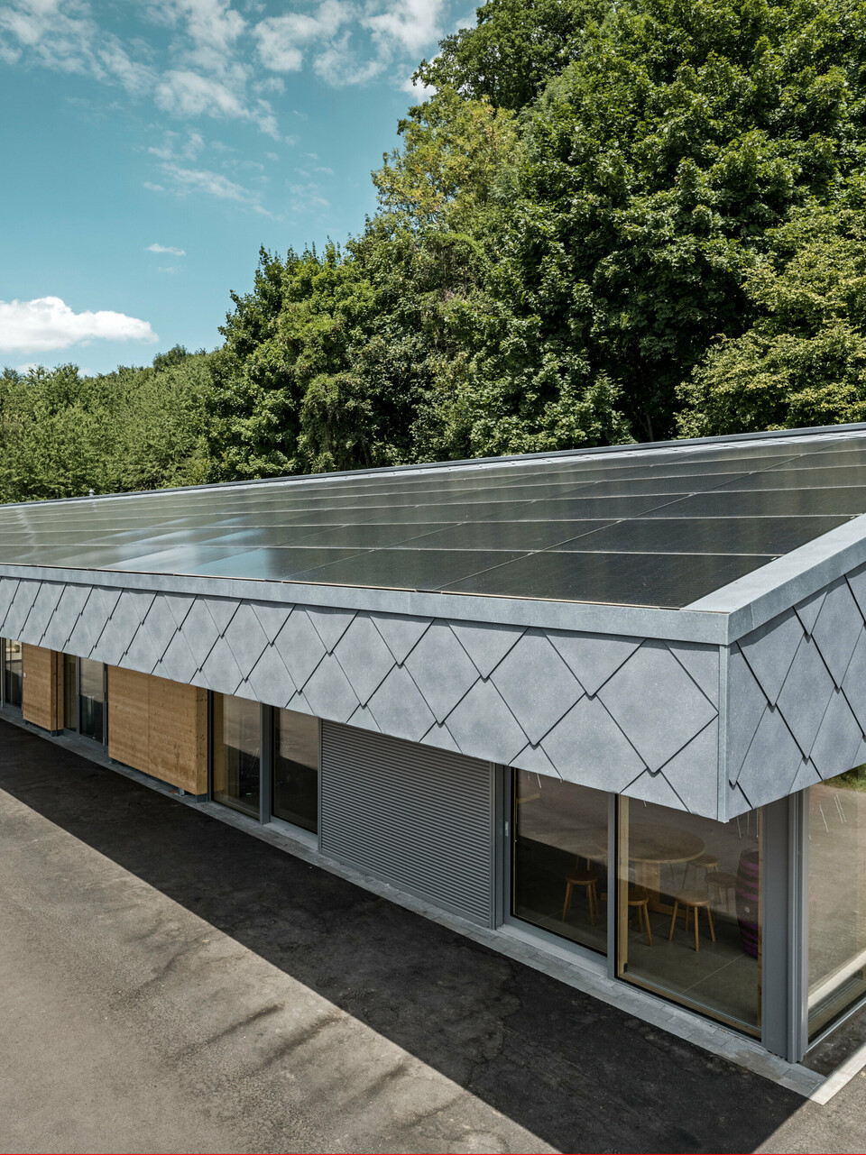 Ansicht von oben auf eine moderne Sportanlage in Prilly, Schweiz. Das Gebäude wurde mit mit langlebigen PREFA Dach- und Wandrauten 44x44 in der Farbe P.10 Steingrau ausgestattet. Die Rauten aus Aluminium sorgen für ein markantes Design, das perfekt mit der großflächig installierten Photovoltaikanlage harmoniert. Diese Kombination zeigt die innovative Verbindung von Funktionalität, Energieeffizienz und ansprechender Ästhetik, die für PREFA Aluminiumprodukte steht. Das Gebäude repräsentiert zukunftsorientiertes Bauen mit einem Fokus auf Nachhaltigkeit und Energieautonomie.
