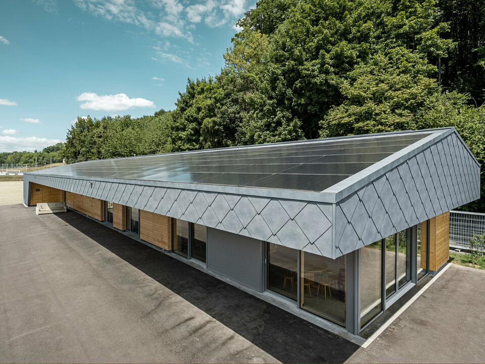 Ansicht von oben auf eine moderne Sportanlage in Prilly, Schweiz. Das Gebäude wurde mit mit langlebigen PREFA Dach- und Wandrauten 44x44 in der Farbe P.10 Steingrau ausgestattet. Die Rauten aus Aluminium sorgen für ein markantes Design, das perfekt mit der großflächig installierten Photovoltaikanlage harmoniert. Diese Kombination zeigt die innovative Verbindung von Funktionalität, Energieeffizienz und ansprechender Ästhetik, die für PREFA Aluminiumprodukte steht. Das Gebäude repräsentiert zukunftsorientiertes Bauen mit einem Fokus auf Nachhaltigkeit und Energieautonomie.