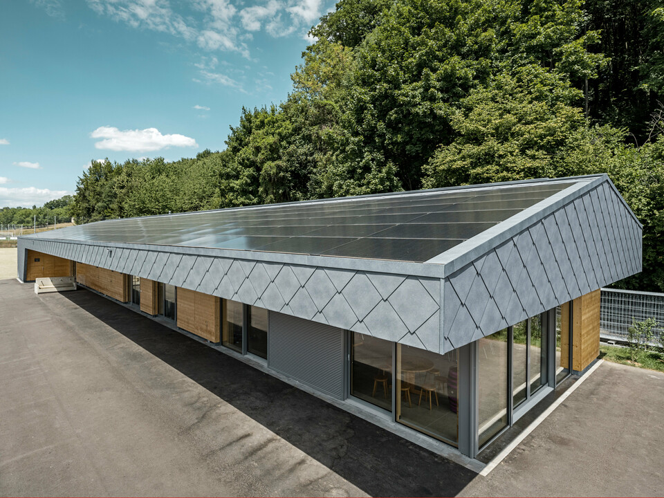 Ansicht von oben auf eine moderne Sportanlage in Prilly, Schweiz. Das Gebäude wurde mit mit langlebigen PREFA Dach- und Wandrauten 44x44 in der Farbe P.10 Steingrau ausgestattet. Die Rauten aus Aluminium sorgen für ein markantes Design, das perfekt mit der großflächig installierten Photovoltaikanlage harmoniert. Diese Kombination zeigt die innovative Verbindung von Funktionalität, Energieeffizienz und ansprechender Ästhetik, die für PREFA Aluminiumprodukte steht. Das Gebäude repräsentiert zukunftsorientiertes Bauen mit einem Fokus auf Nachhaltigkeit und Energieautonomie.
