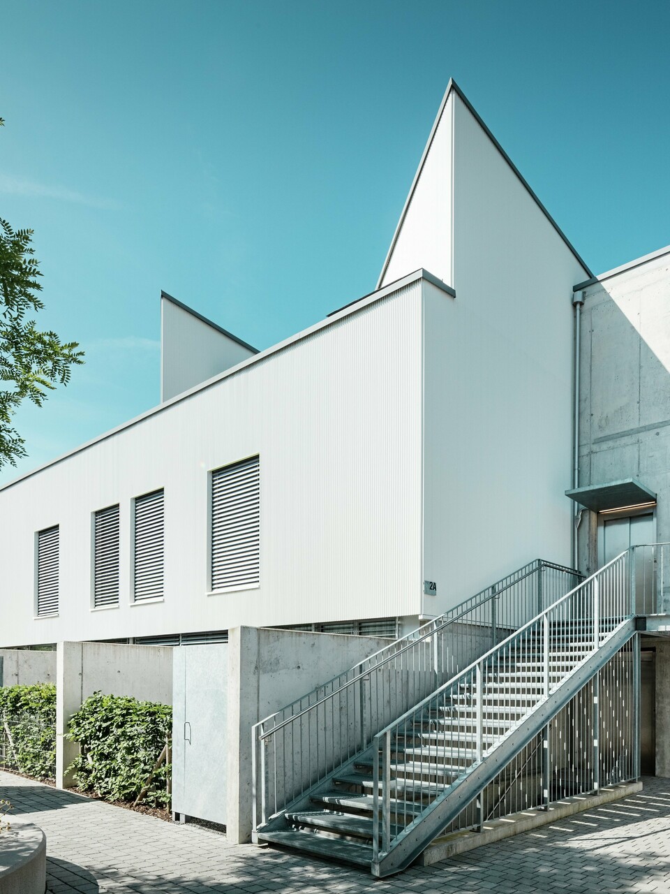 Ensemble moderner Tiny Homes mit markanten Spitzdächern, verkleidet mit dem PREFA Zackenprofil in der Farbe P.10 Reinweiß. Die schlichte, klare Fassadengestaltung harmoniert mit den Sichtbetonelementen und der umlaufenden Stahlkonstruktion. Die langlebige PREFA Aluminiumfassade schützt zuverlässig vor Witterung und sorgt für eine wartungsarme, ästhetisch reduzierte Optik.