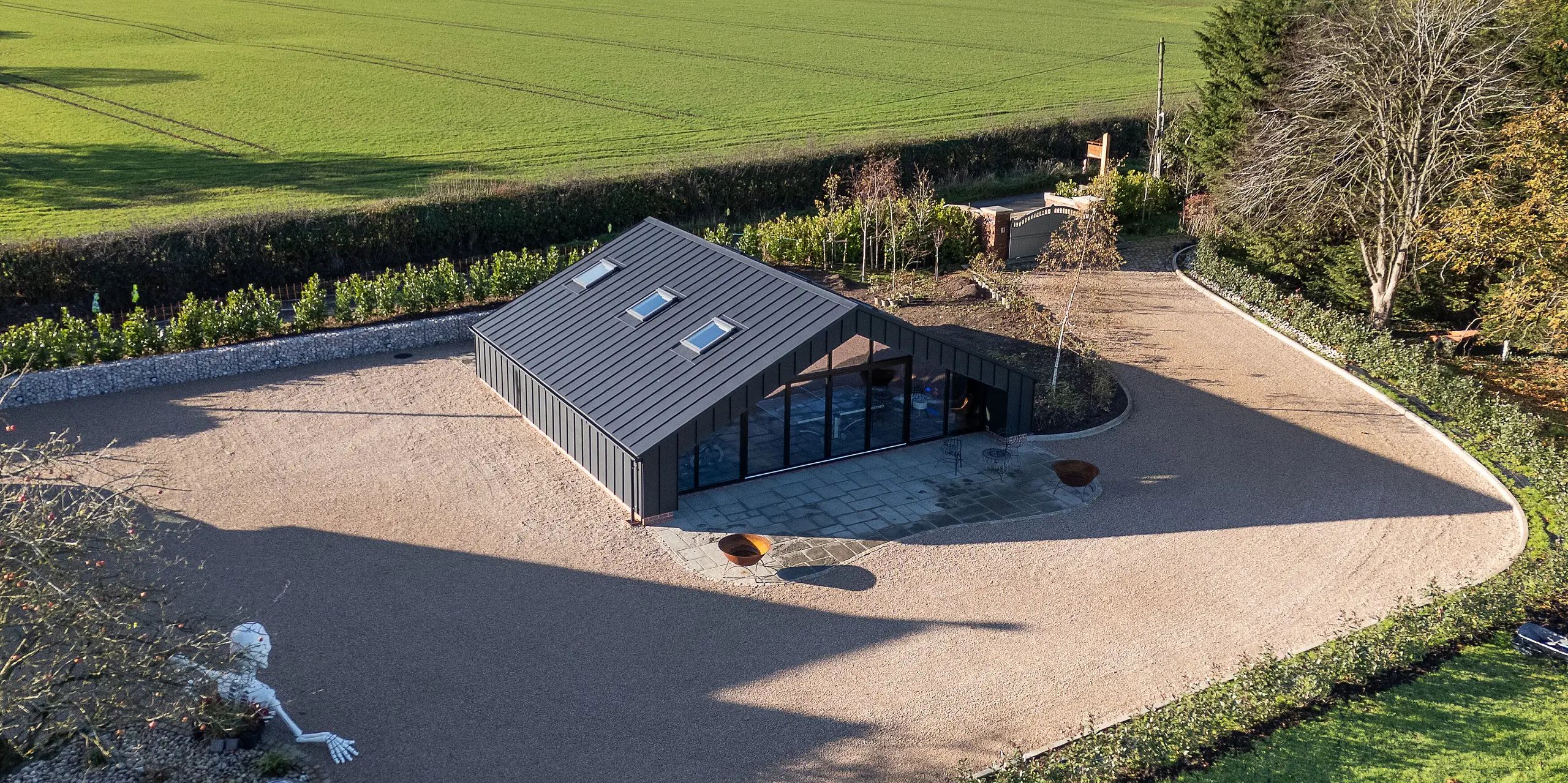 [Translate to Français:] Weitläufige Vogelperspektive auf das moderne Freizeitgebäude mit vollständig verkleideter PREFA Aluminiumfassade und PREFALZ Dachflächen in P.10 Schwarz. Die präzisen Stehfalzlinien, die markante Dachgeometrie und die großflächige Glasfront setzen starke architektonische Akzente. Die robuste, witterungsbeständige Aluminiumhülle fügt sich harmonisch in die offene Landschaft ein.