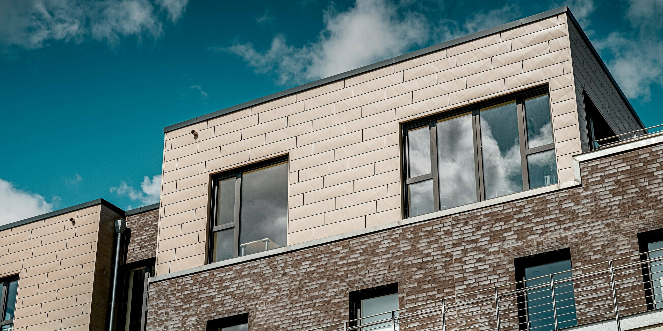 Die moderne Wohnanlage „Hafen zur Freiheit“ legt den Fokus auf das elegante Staffelgeschoss, das mit PREFA Siding.X in einem warmen Bronzeton verkleidet ist. Die charakteristische, reliefartige Struktur der Aluminiumpaneele bildet einen spannenden Kontrast zur dunklen Klinkerfassade darunter. Großzügige Fensterflächen spiegeln den Himmel wider und unterstreichen den hochwertigen, lichtdurchfluteten Charakter des Gebäudes. Die robusten und witterungsbeständigen PREFA-Fassadenelemente sind in der küstennahen Lage besonders langlebig und geben dem Ensemble seine markante, zeitgemäße Optik.