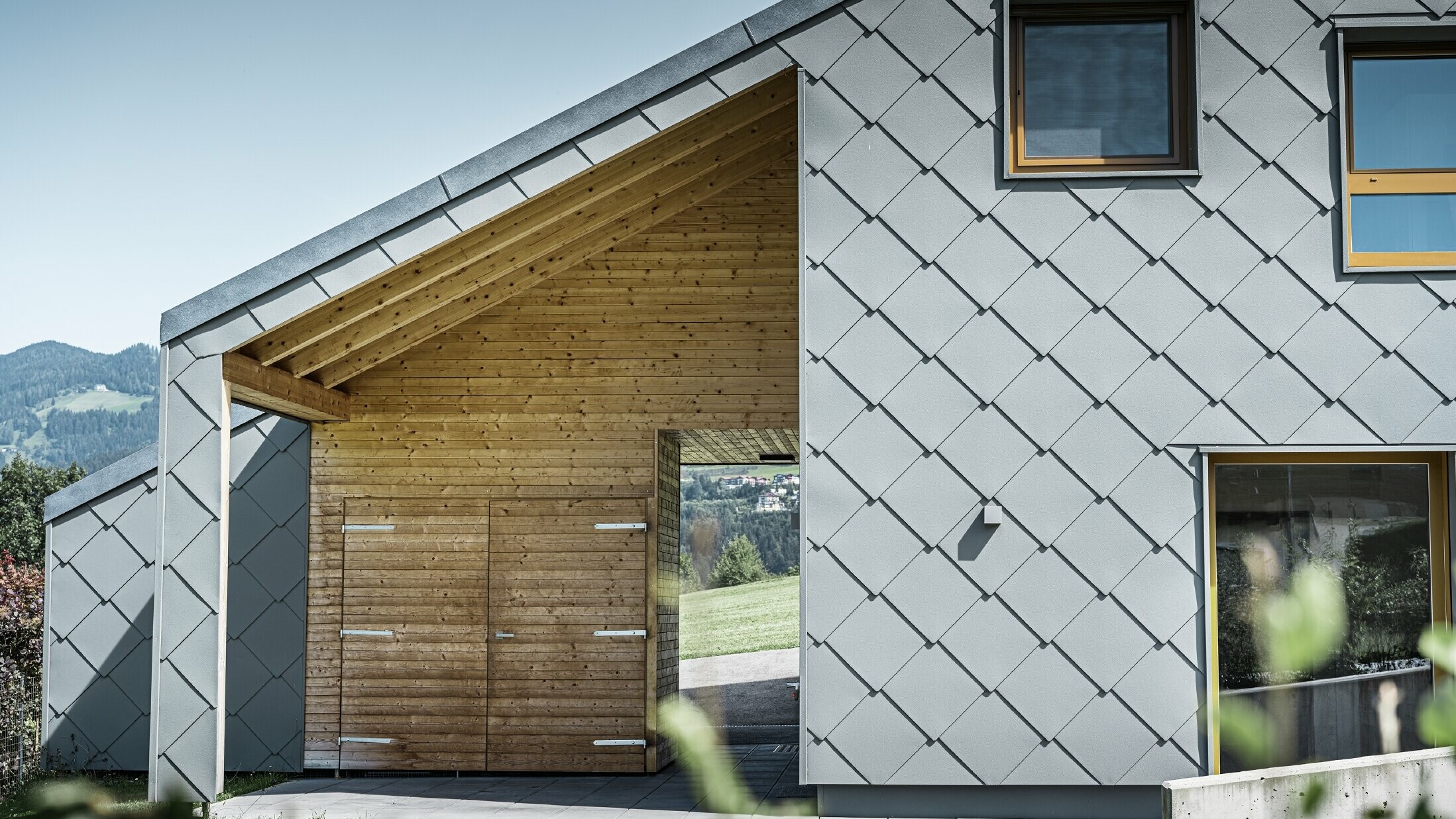 Der überdachte Eingangsbereich ist mit waagrechter Holzverkleidung gemacht, der Rest der Fassade ist mit der großen PREFA Wandraute aus Aluminium in Hellgrau verkleidet